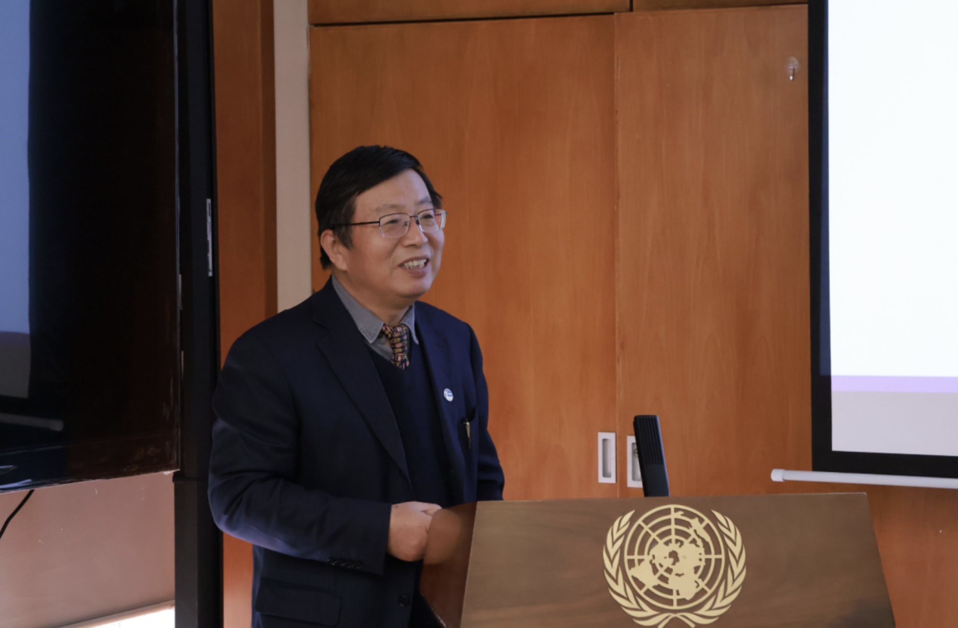 Presenter in a dark blazer at a wooden lectern with microphone and seal emblem.