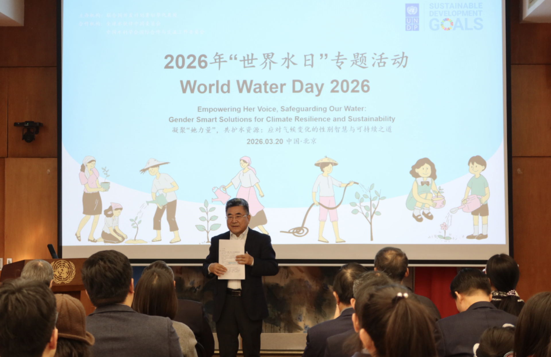 Photo: Speaker on stage at World Water Day 2026; audience watches a screen with cartoon kids.