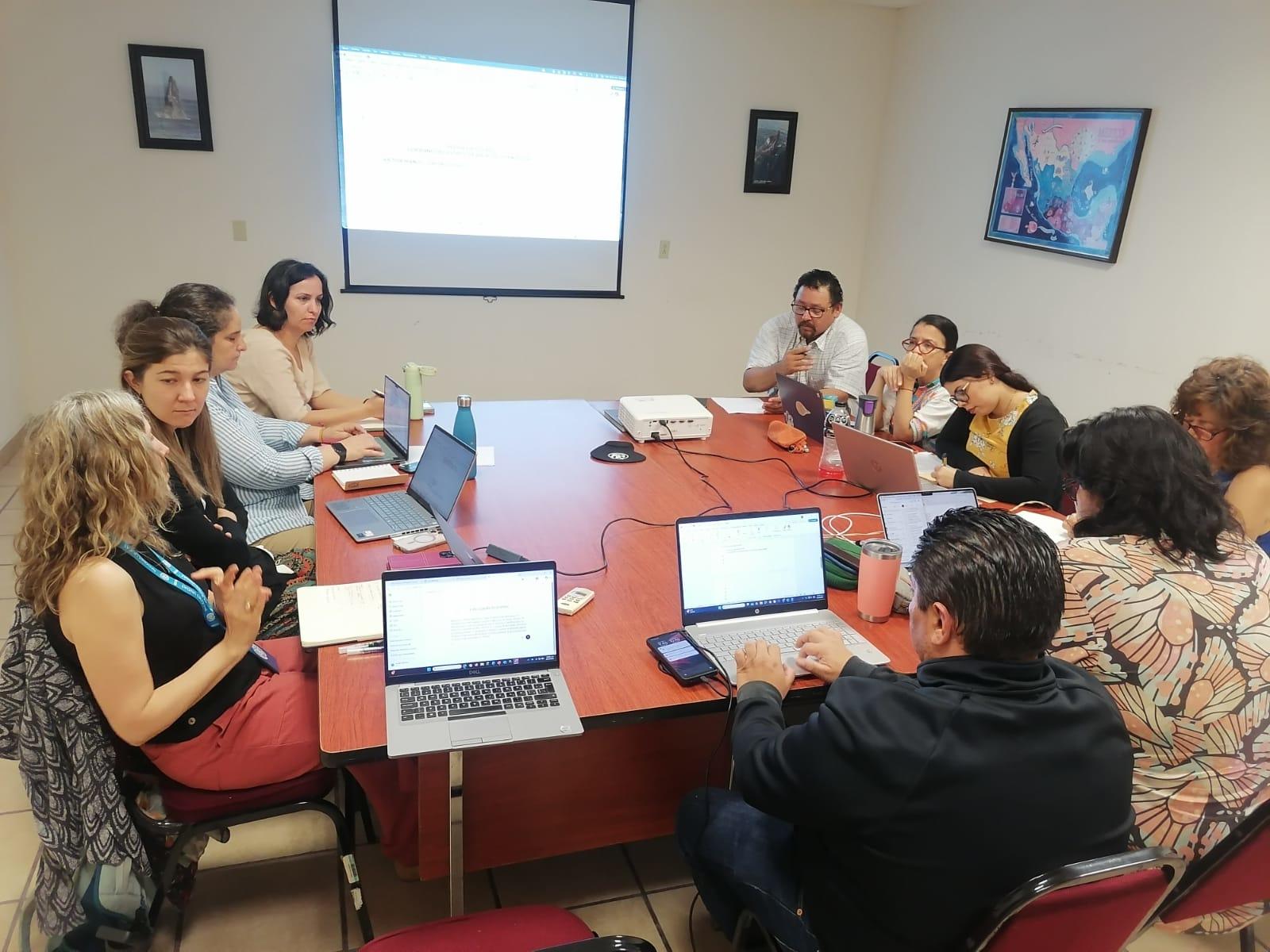 Group of adults in a meeting around a red conference table with laptops; projector screen at front.