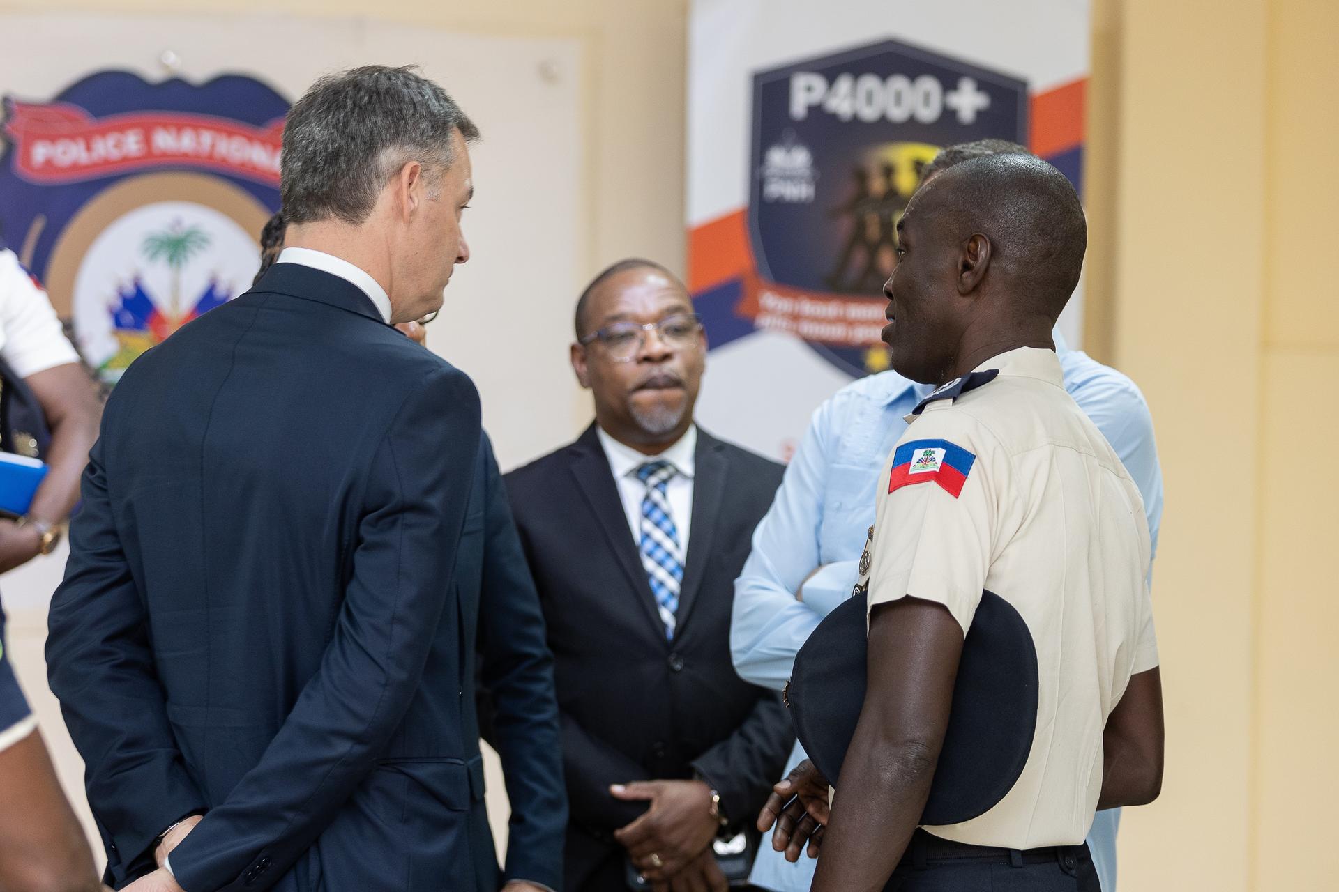 Three men in suits shake hands with a uniformed officer in a hallway.