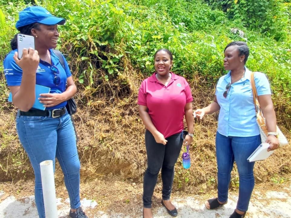 Three people on a dirt path with greenery; a woman in a blue cap speaks, others listen.