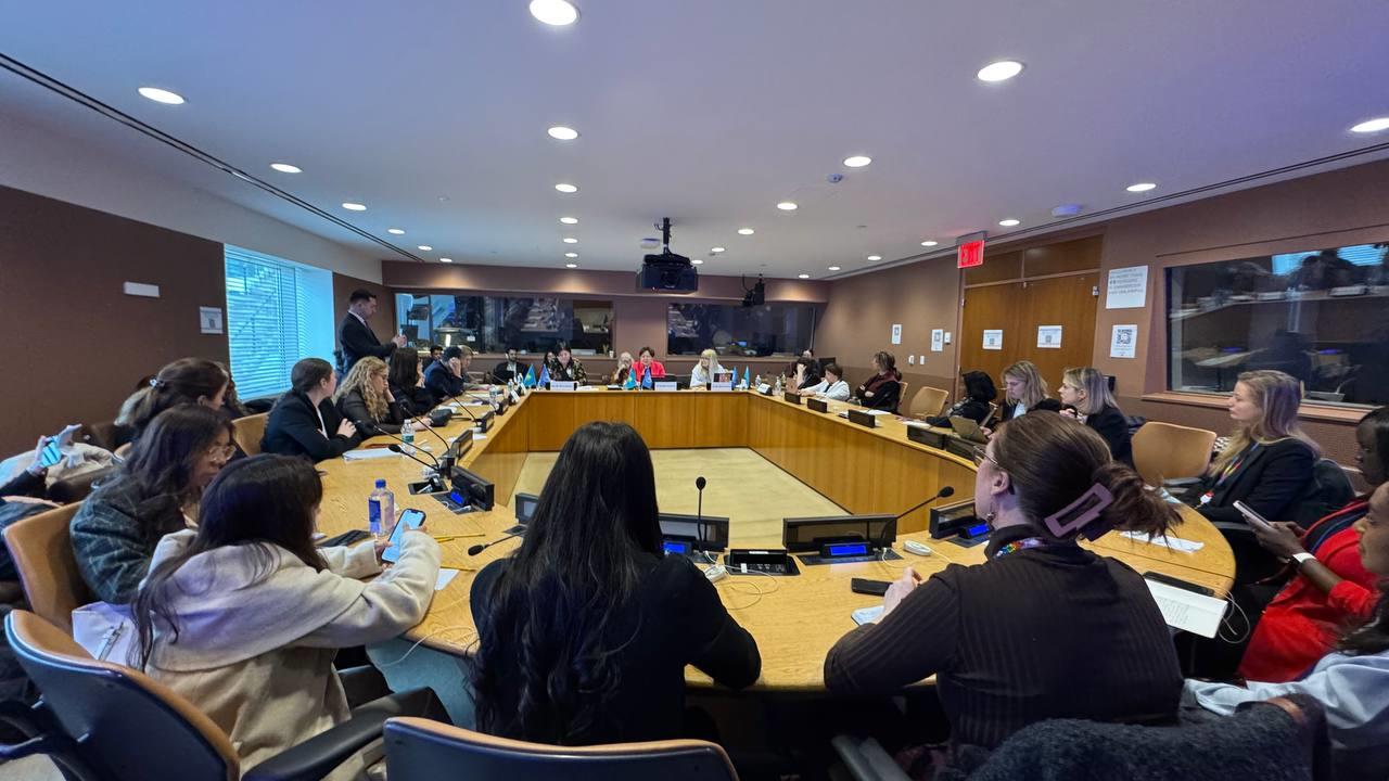 Large conference room with a long table, many attendees seated around it, a speaker at the far end.
