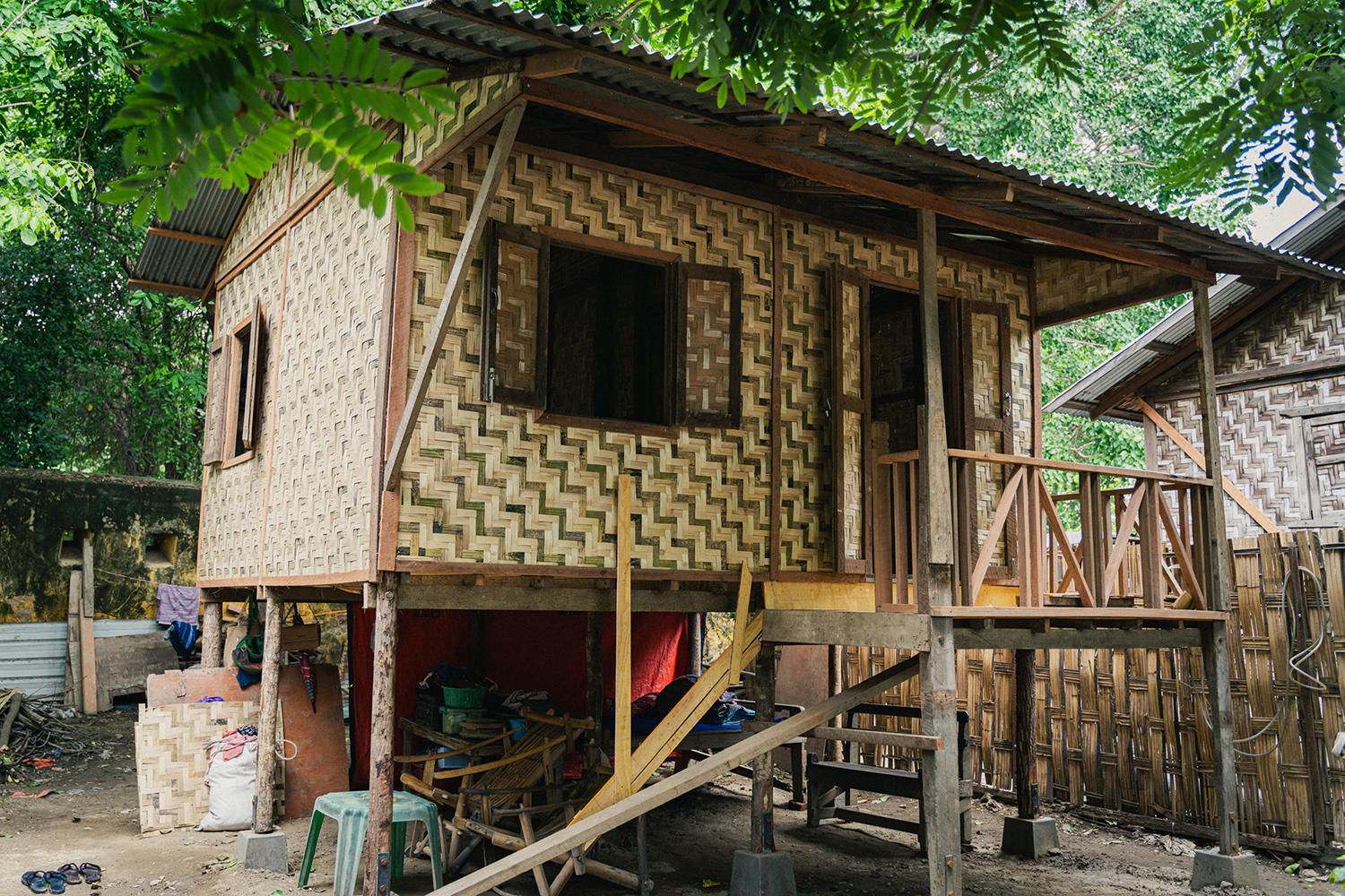 Woven-bamboo stilt house with a wooden porch; a person on the porch among leafy trees.