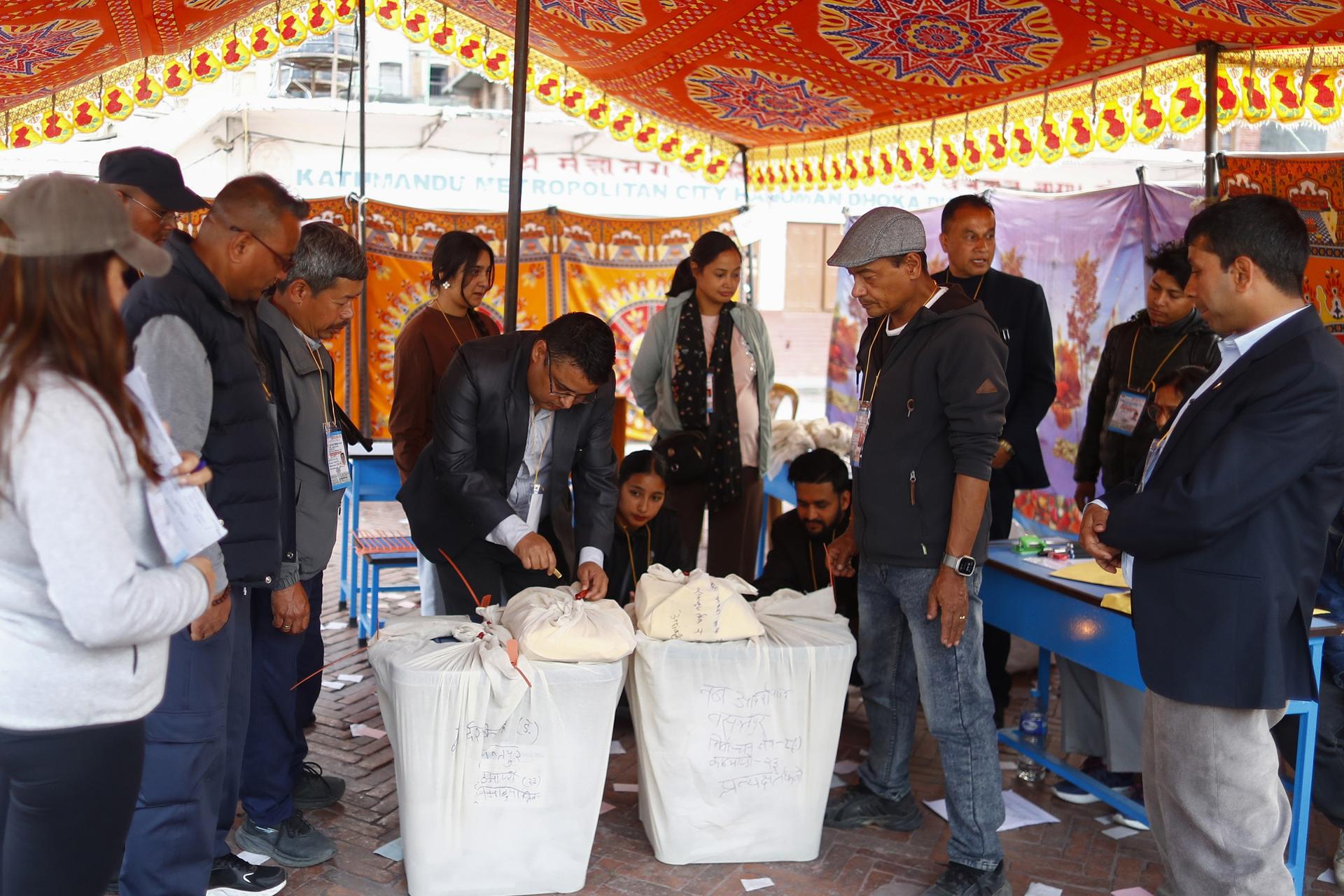 Photograph of people under an orange-patterned tent at an outdoor event, gathered around three white bags.