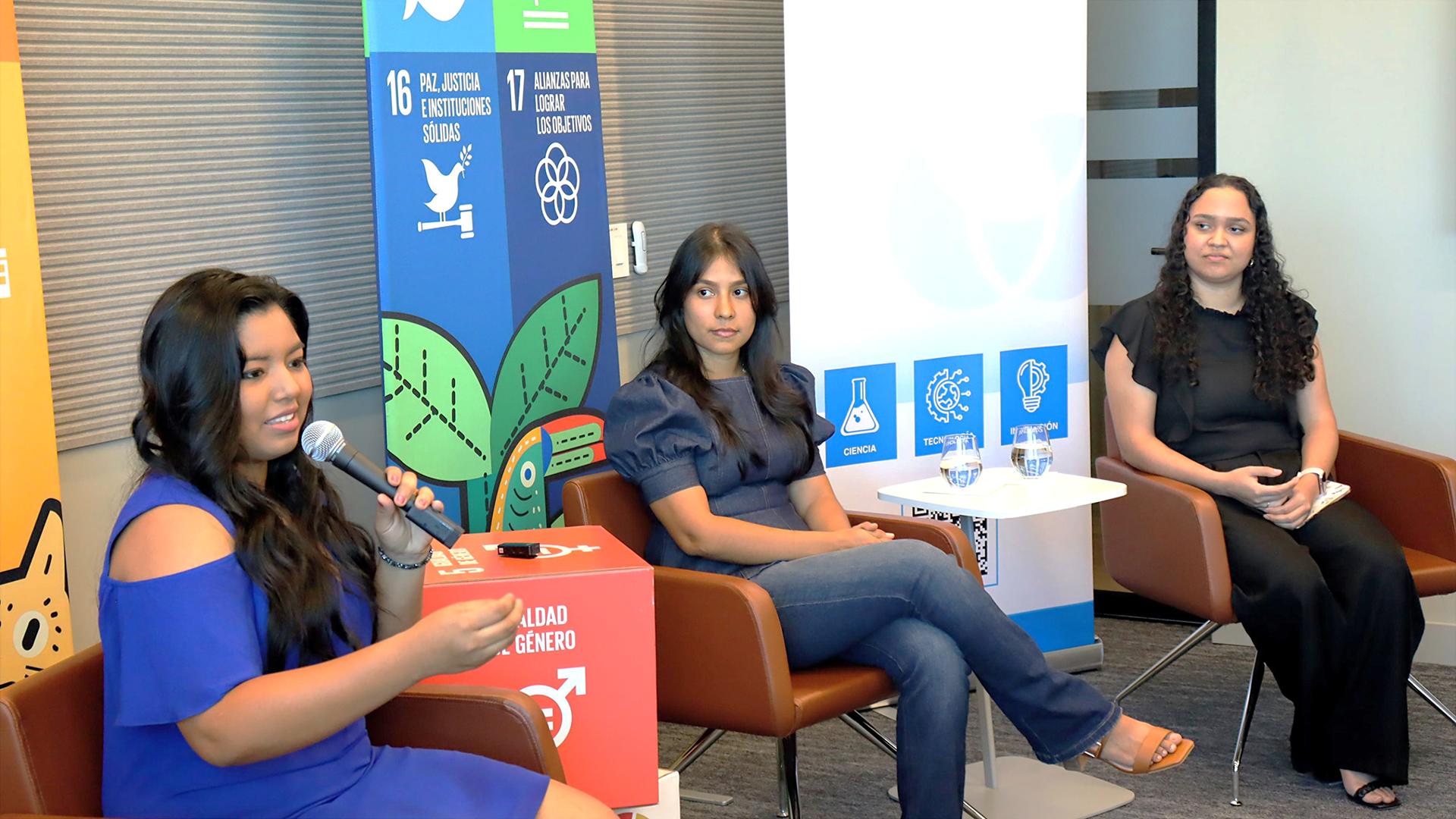 Tres mujeres panelistas sentadas en sillas durante una discusión, con pancartas coloridas detrás de ellas.