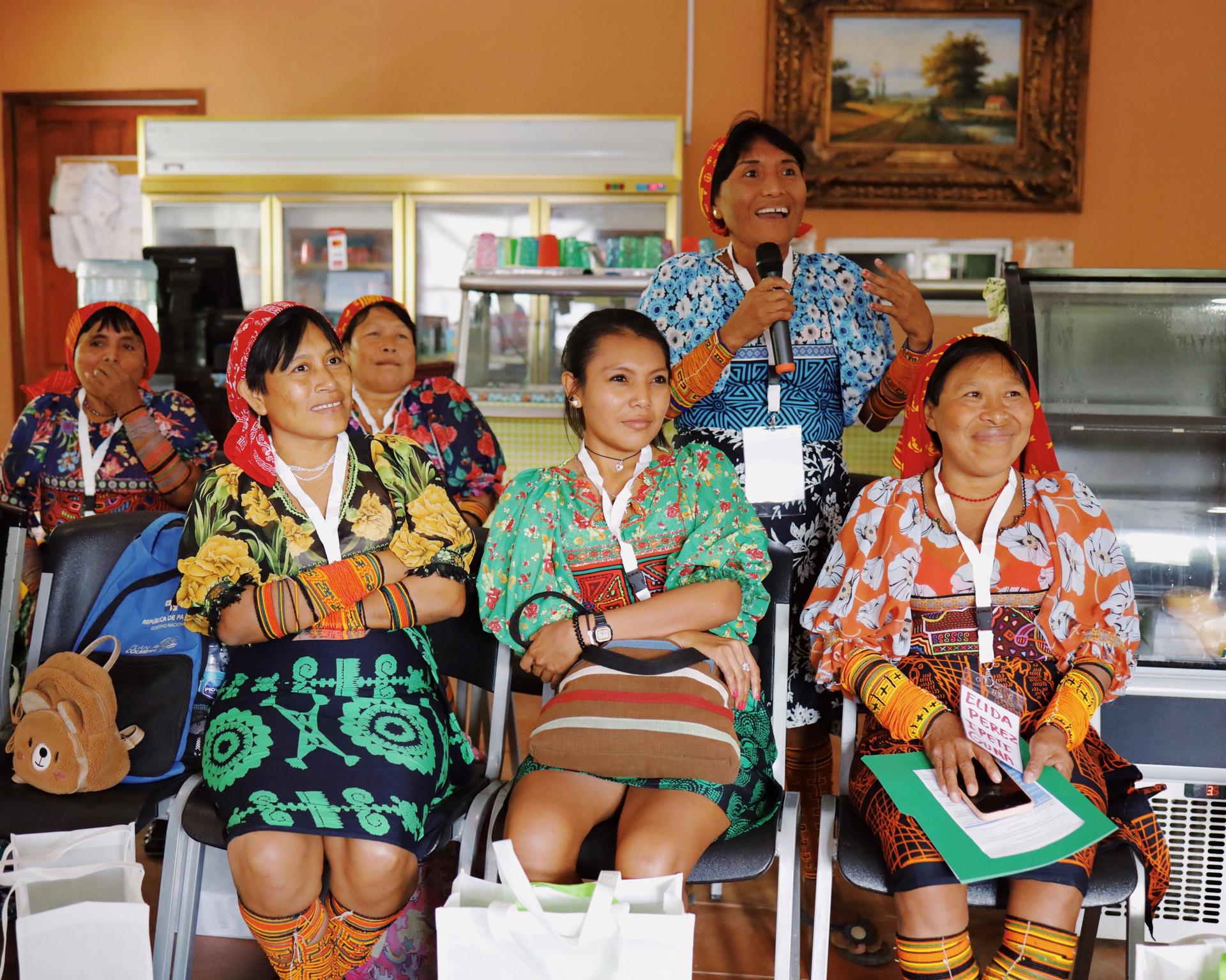 Un grupo de mujeres indígenas están sentadas y visten vestidos coloridos