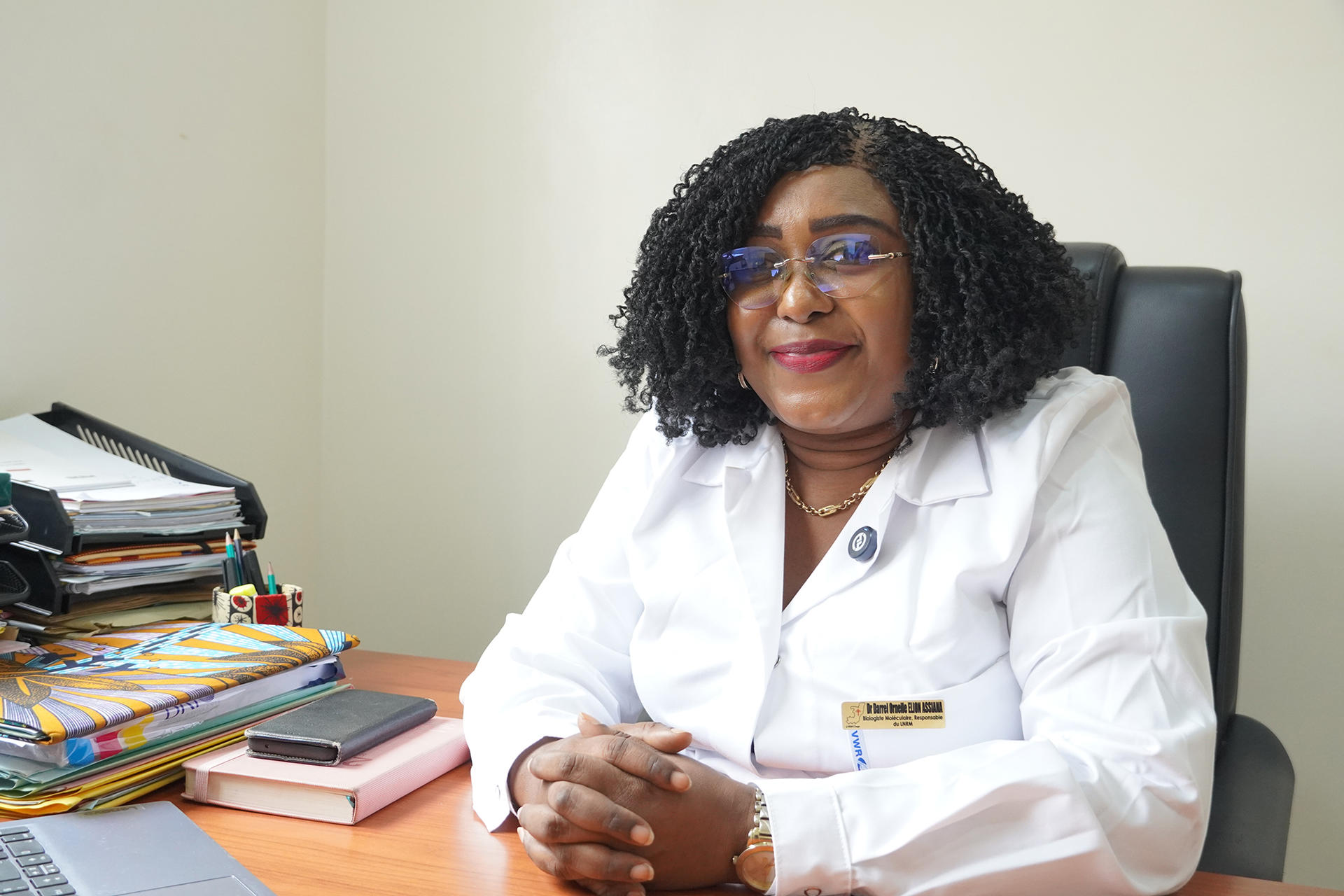 Doctor in white lab coat with her folded hands at a desk; stacks of folders and books nearby.