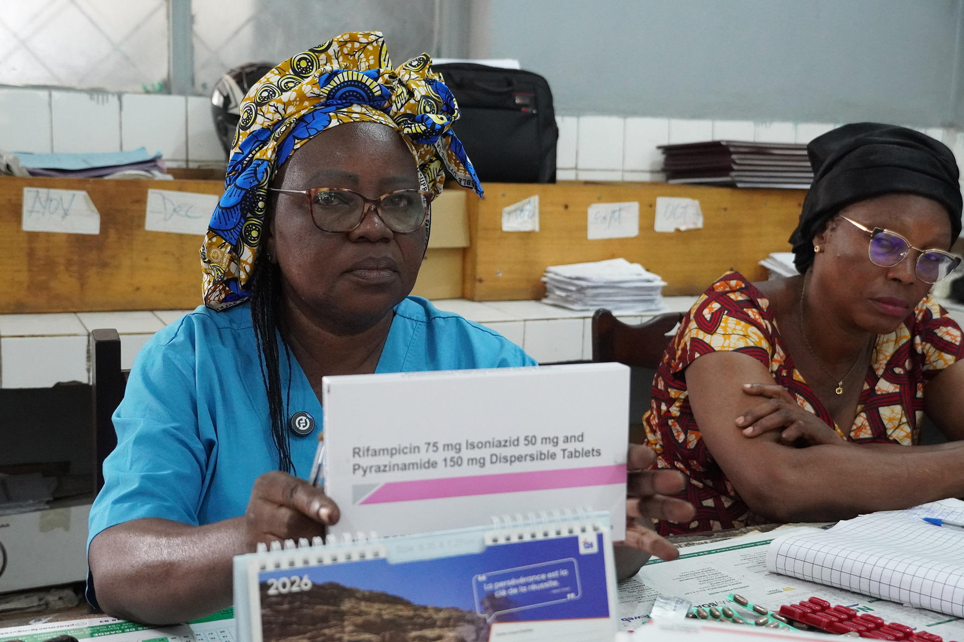 Two women sit at a table with papers and a sign, one wears blue shirt.