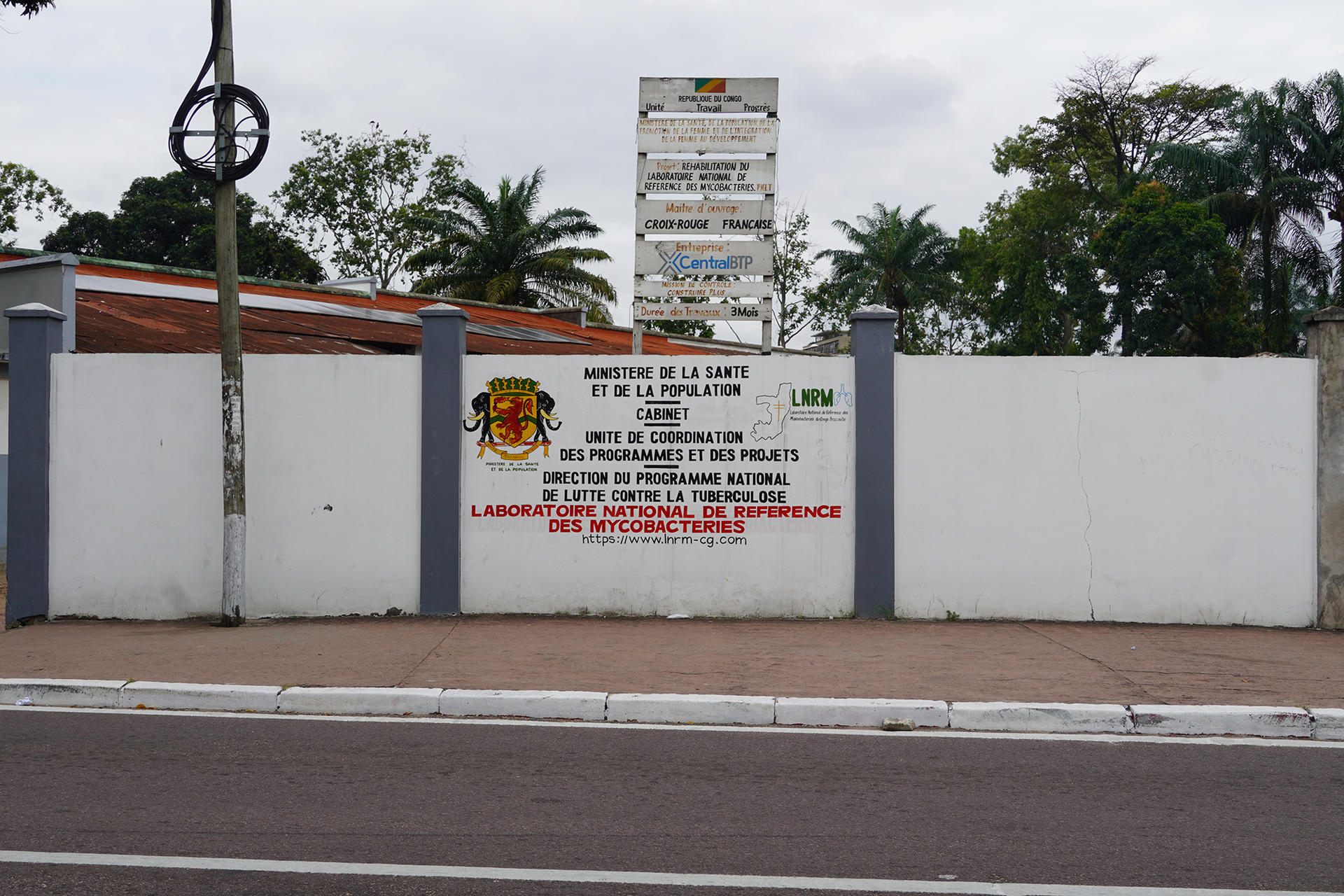 White wall of a laboratory with a large sign and a smaller sign along a street; palm trees and cloudy sky behind.