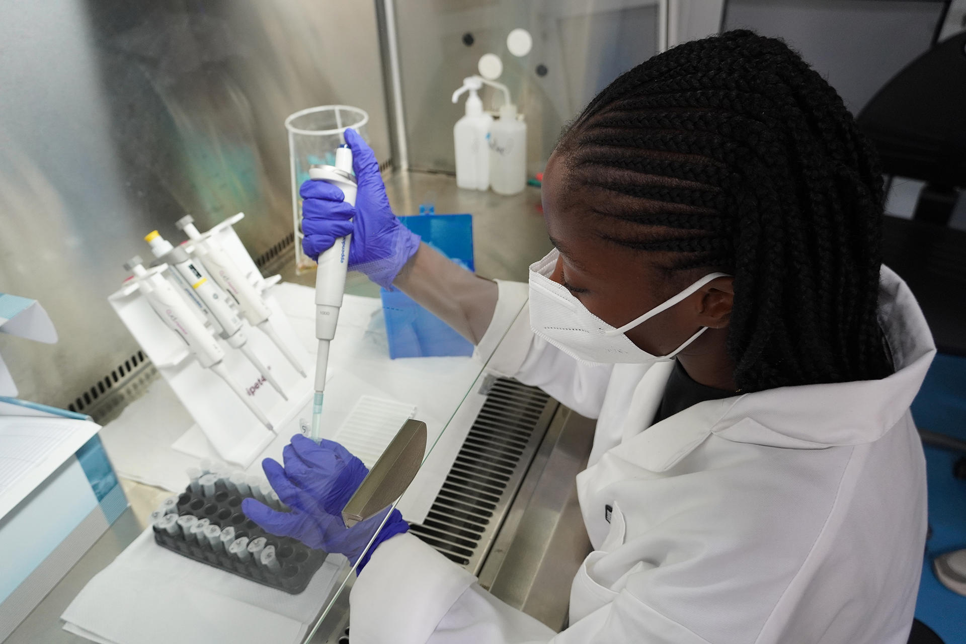 Female scientist in a lab coat, purple gloves, and mask pipetting at a table.