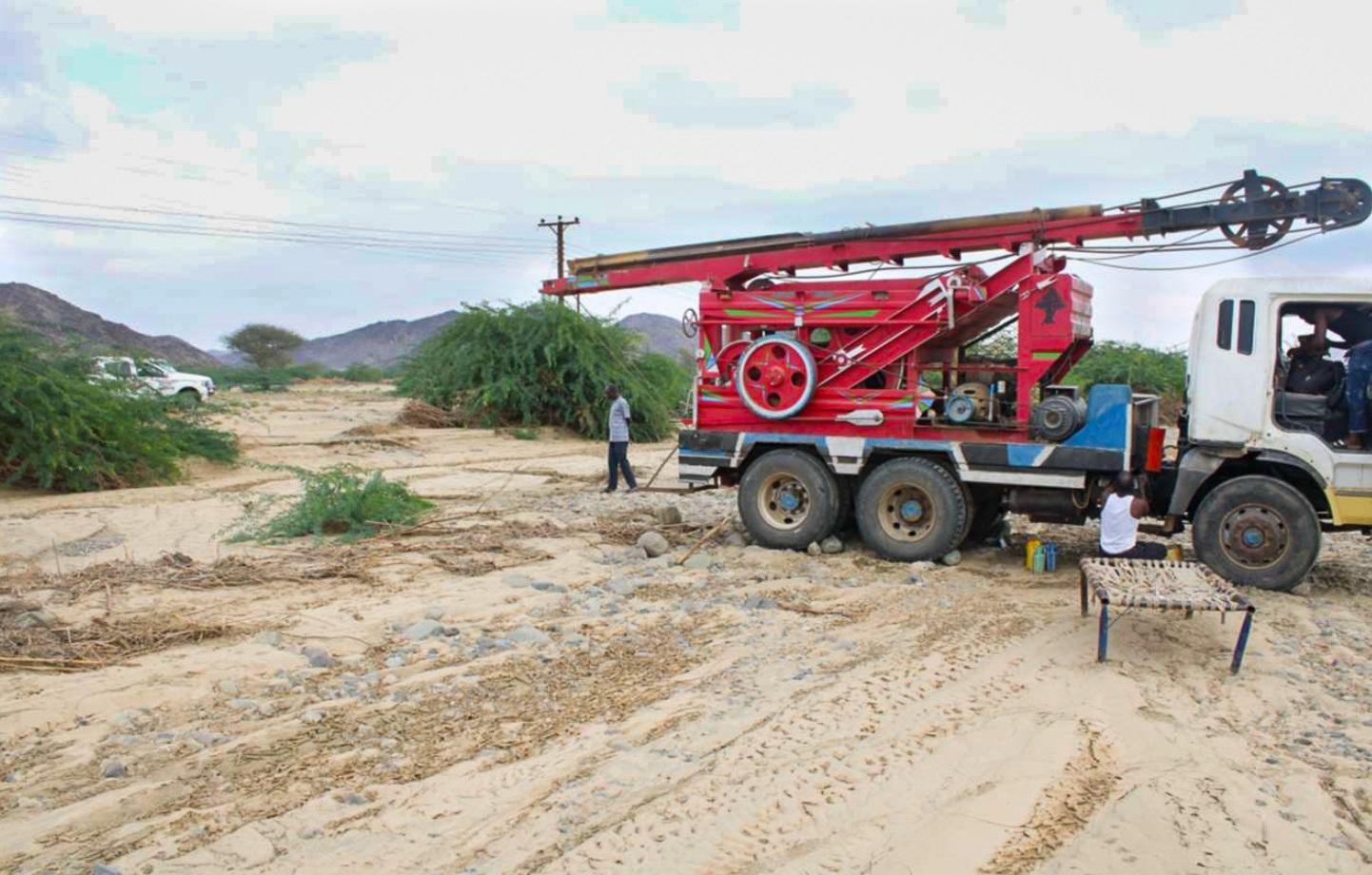 White truck with red crane rig on a sandy construction site, a person nearby.