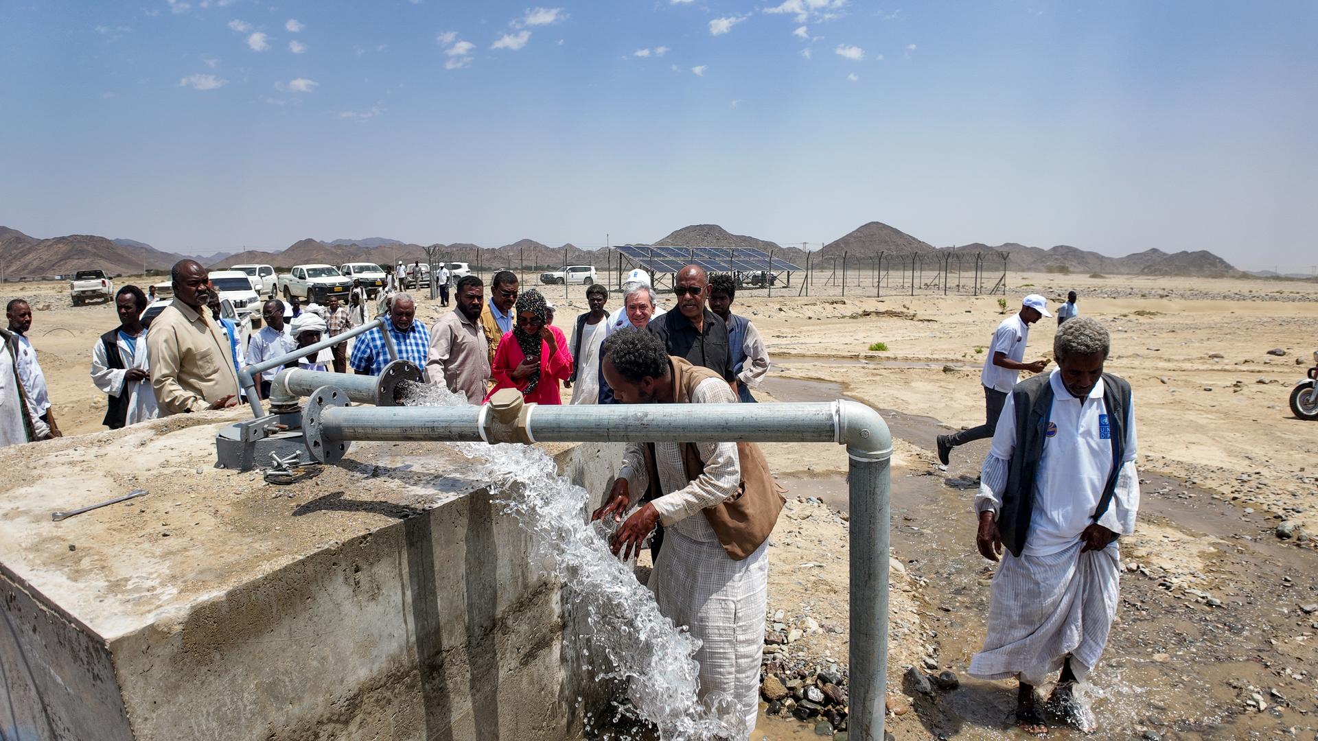 Crowd of travelers near a low concrete barrier in a desert, with blue sky and distant hills.