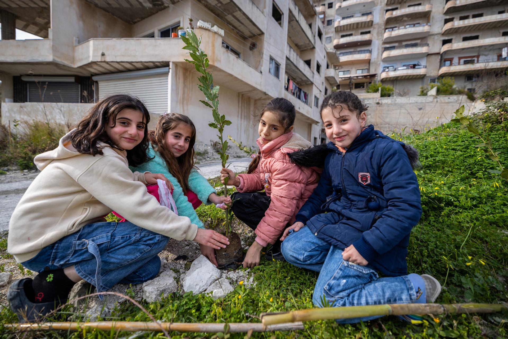 Four kids sit on grass in an outdoor courtyard around a small sapling, smiling.