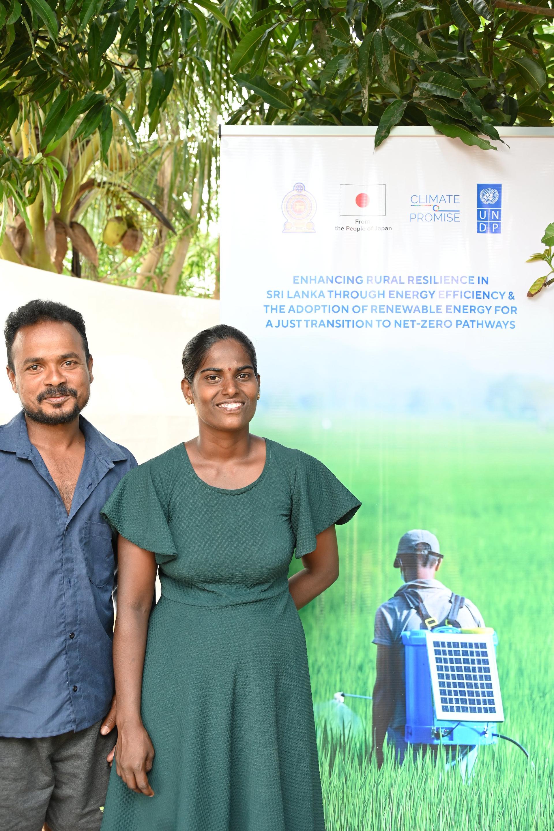 Two people pose outdoors in front of a large poster featuring a solar panel image.