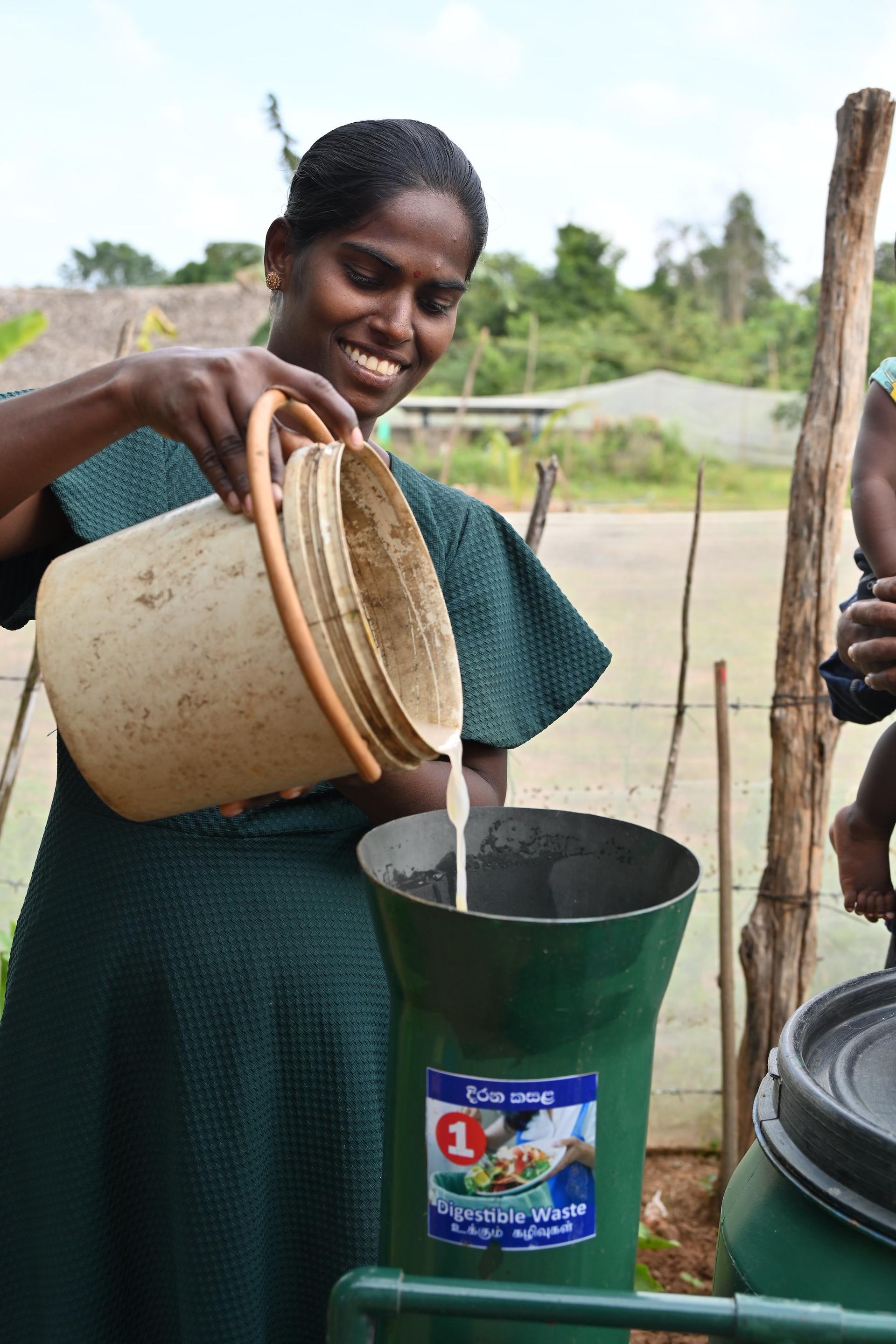 Person in green pours water from a clay pot into a black bucket outdoors in a rural setting.