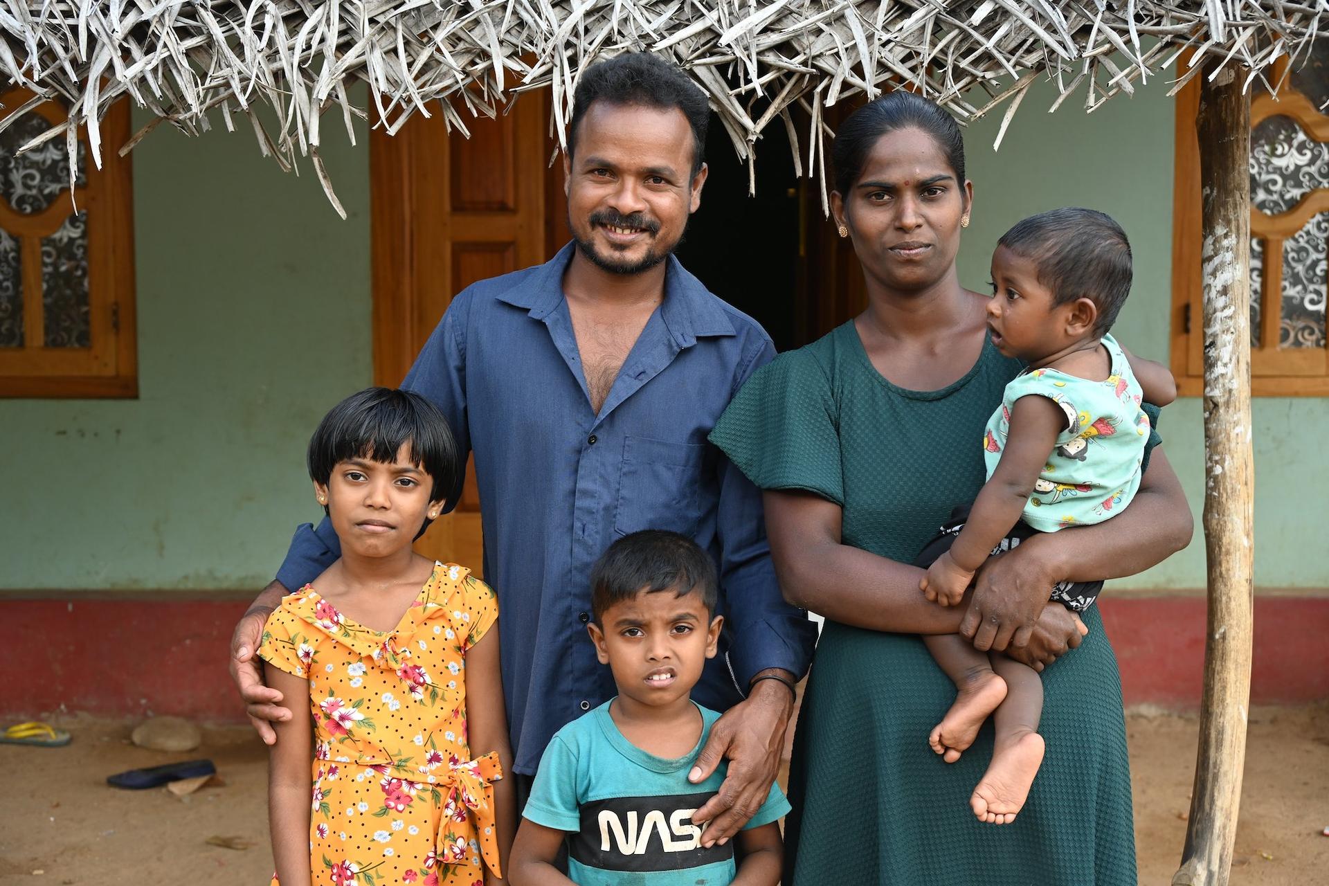 Family of five stands outside a small house with a thatched roof; children wear bright clothes.