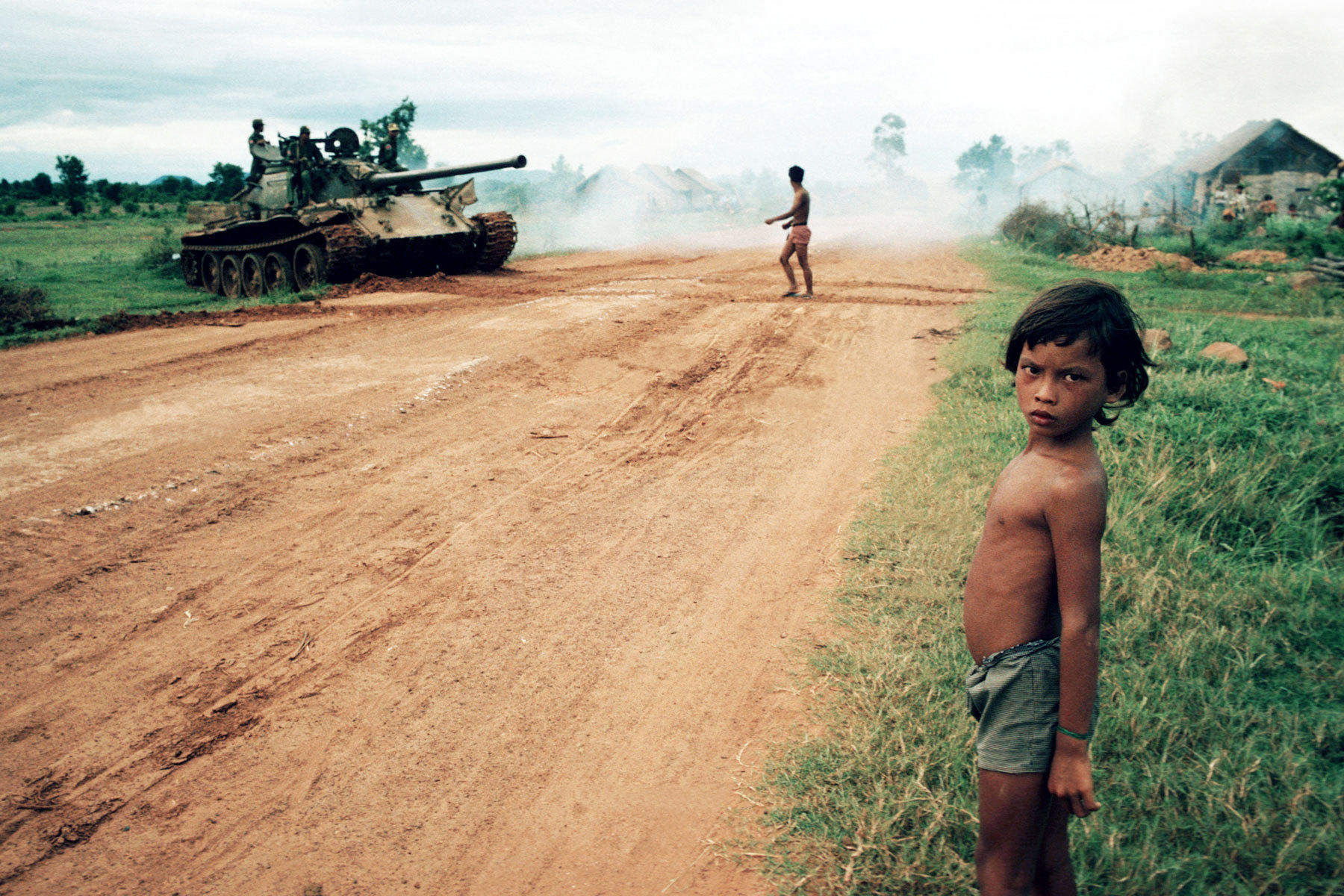 Dusty rural road with tanks on the left; a shirtless boy stands on the grassy verge.