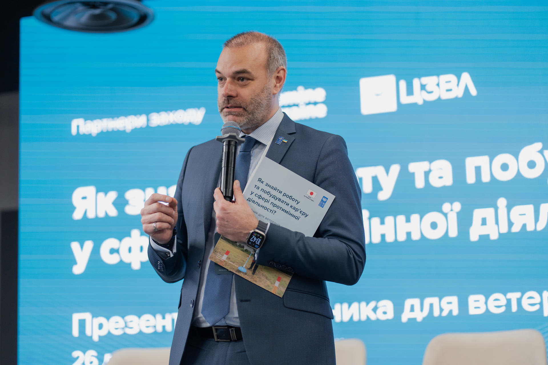 Speaker in a dark suit on a blue stage, holding a microphone and notes.