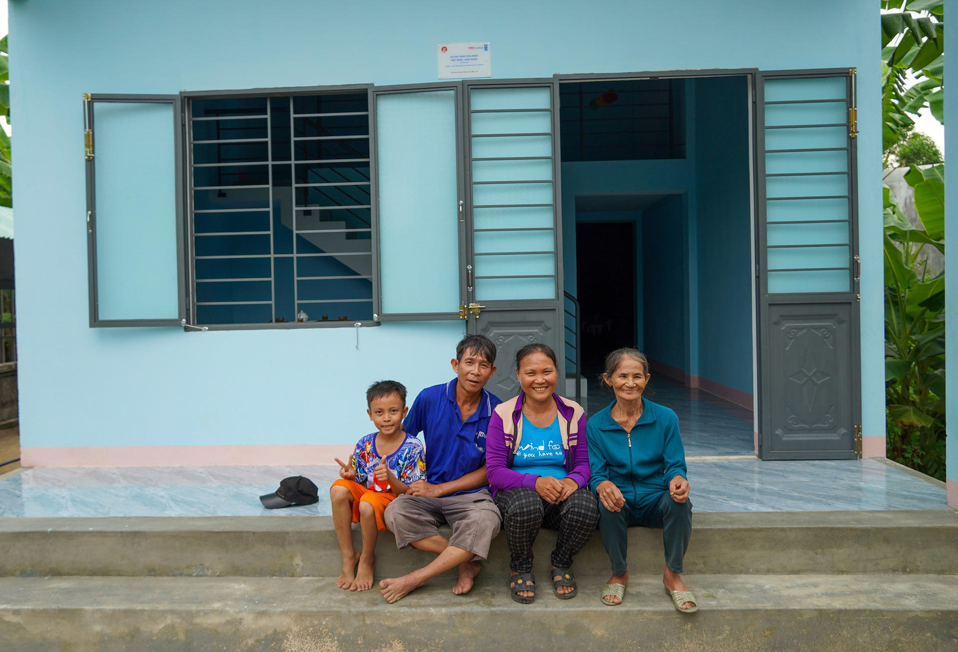 Smiling family sits on concrete steps in front of a blue house with open shutters.