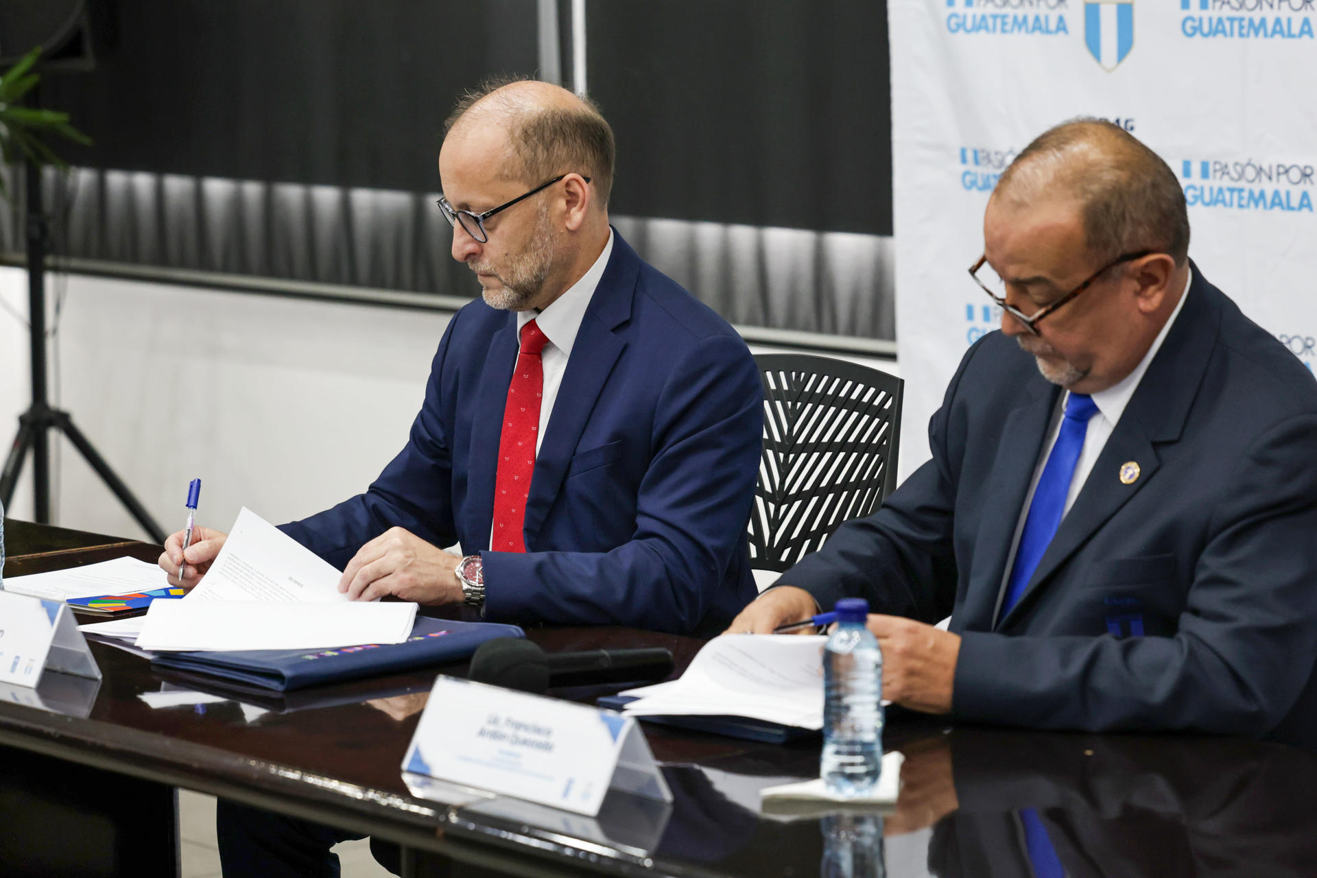 Two men in business suits sign documents at a conference table.