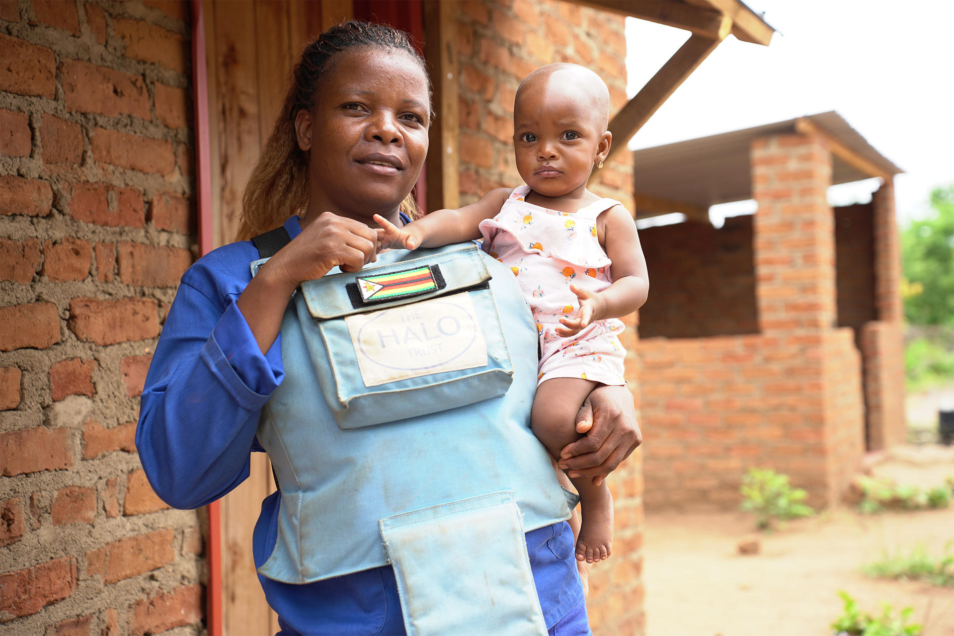 Woman de-miner in blue holds a baby in pink near a brick building outdoors.