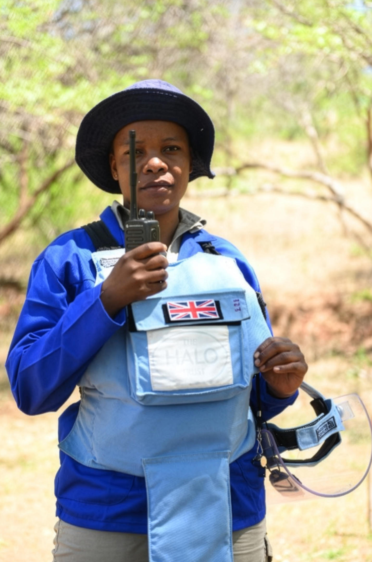 Person in blue shirt and light-blue vest with Union Jack patch outdoors, holding a smartphone.