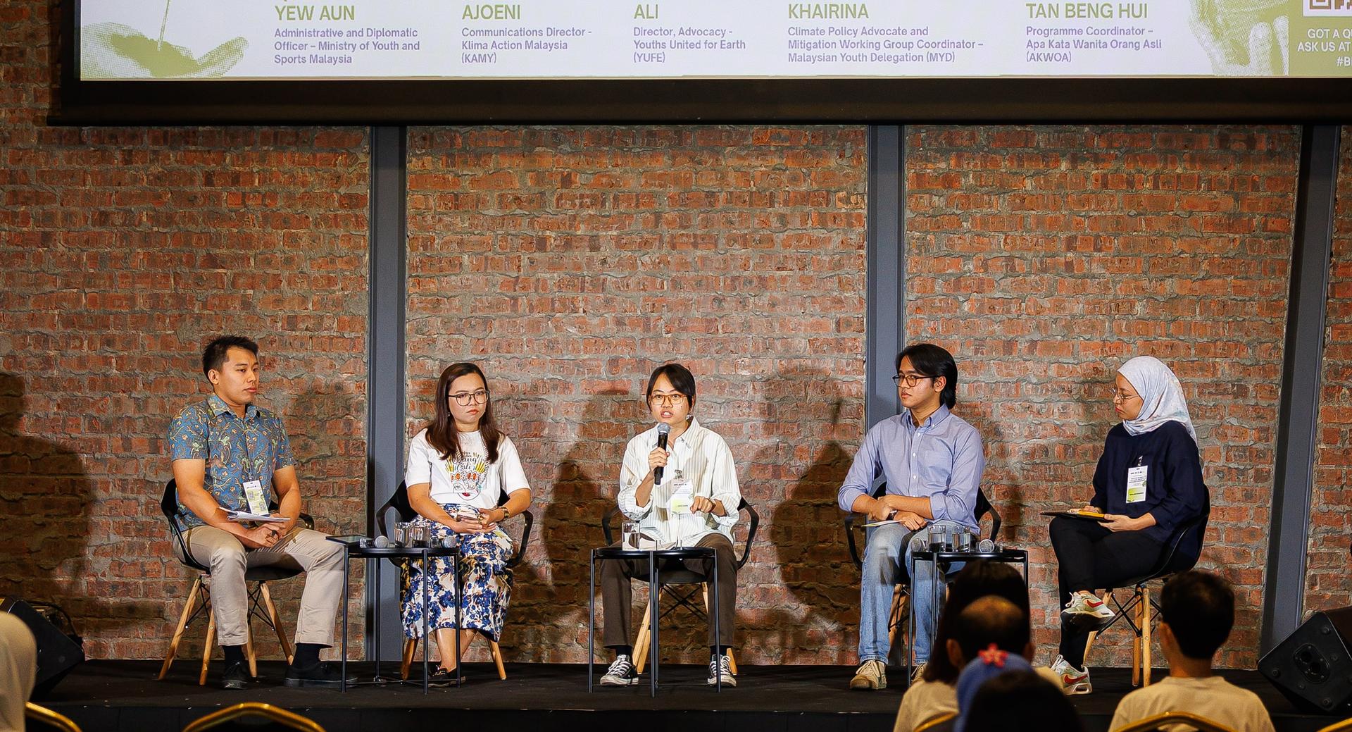 Six panelists seated on a stage with a brick wall backdrop; audience in the foreground.