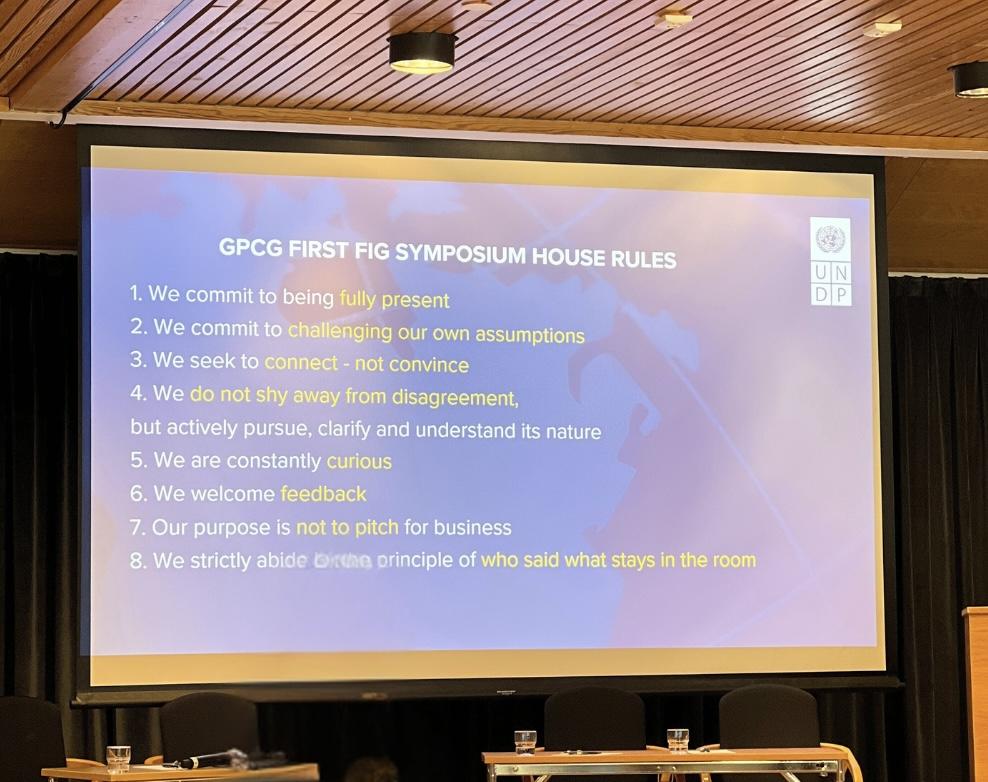 Projected slide listing house rules for the symposium, blue background, auditorium stage.