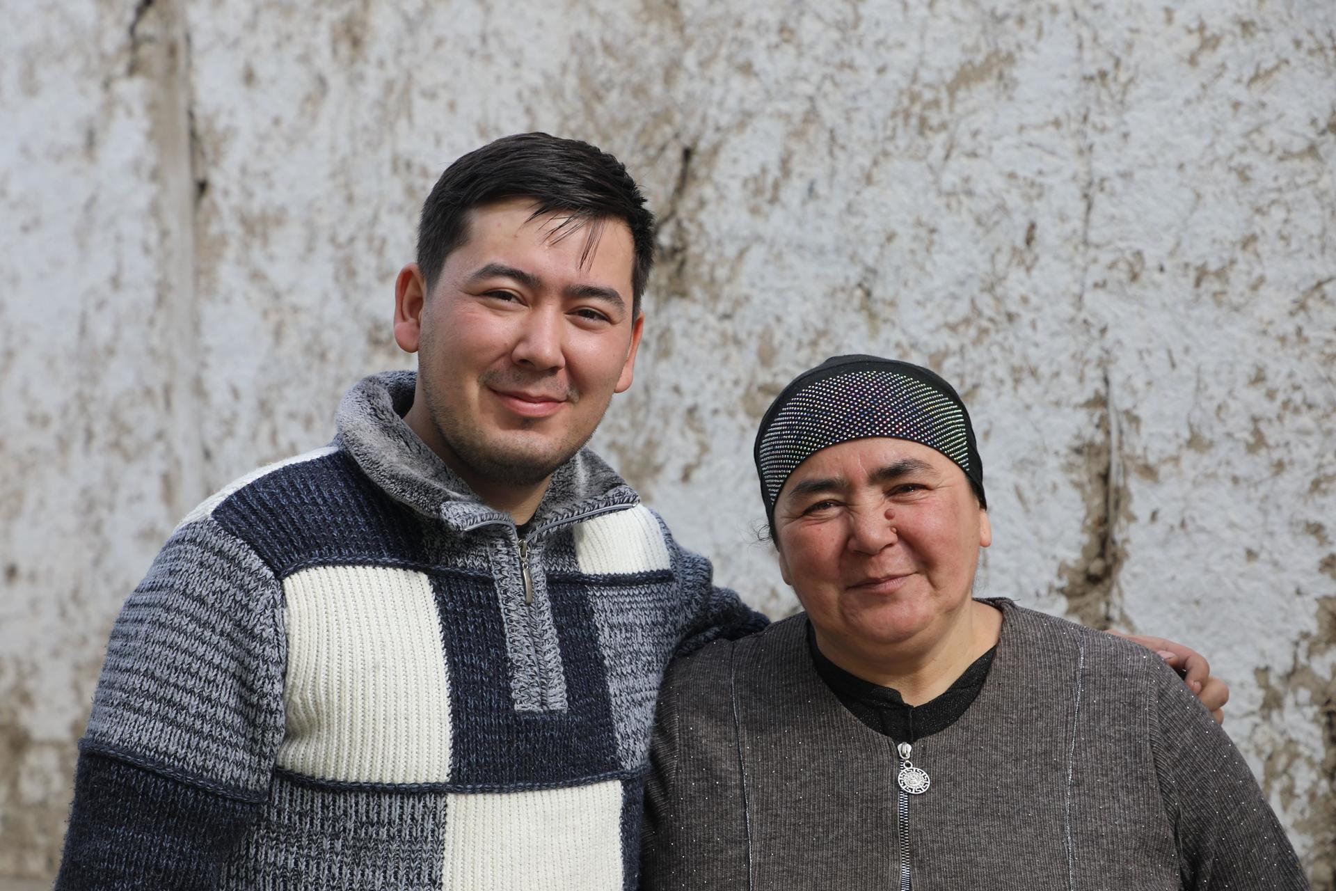 Two men stand in front of a rough stone wall; one in a striped sweater, the other in a dark top.