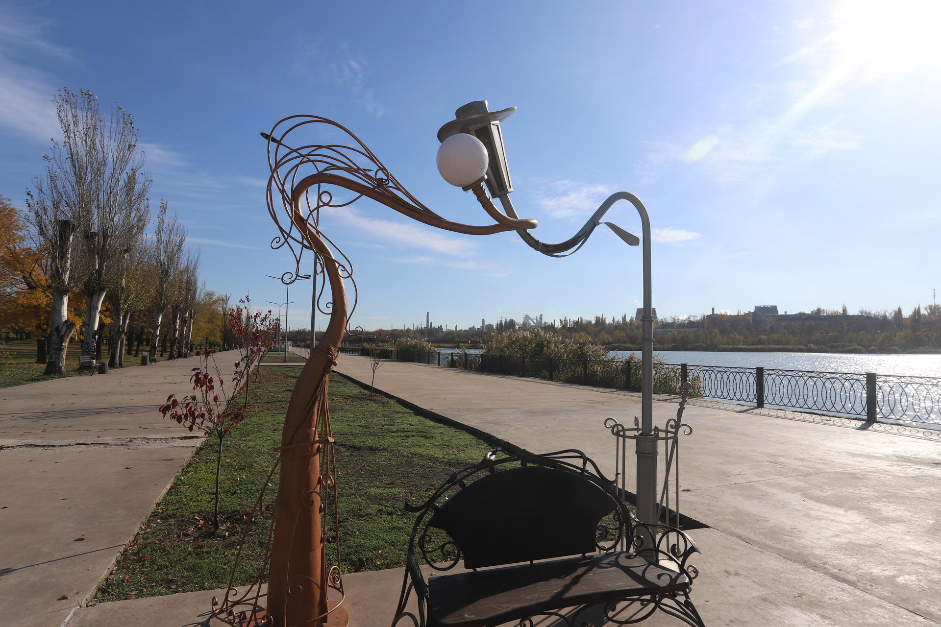Photograph of a metal bird sculpture perched on a curved branch beside a waterfront promenade.