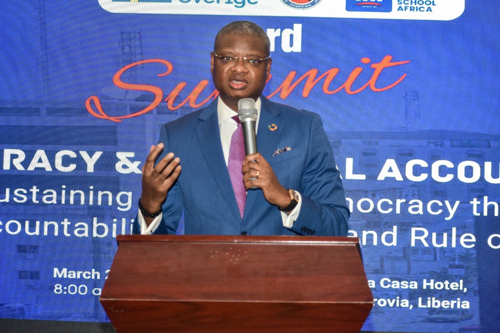 Speaker in a dark suit at a wooden podium, holding a microphone on a blue summit backdrop.