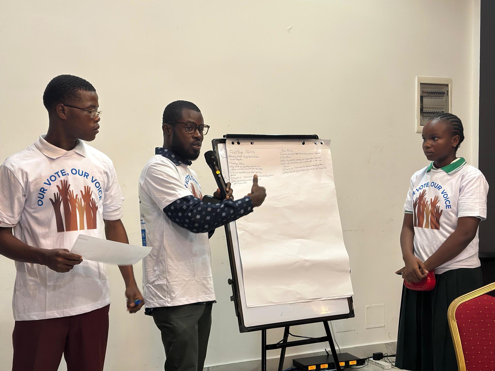 Three men in white T-shirts stand in room during a presentation; one points to a blank flip chart.