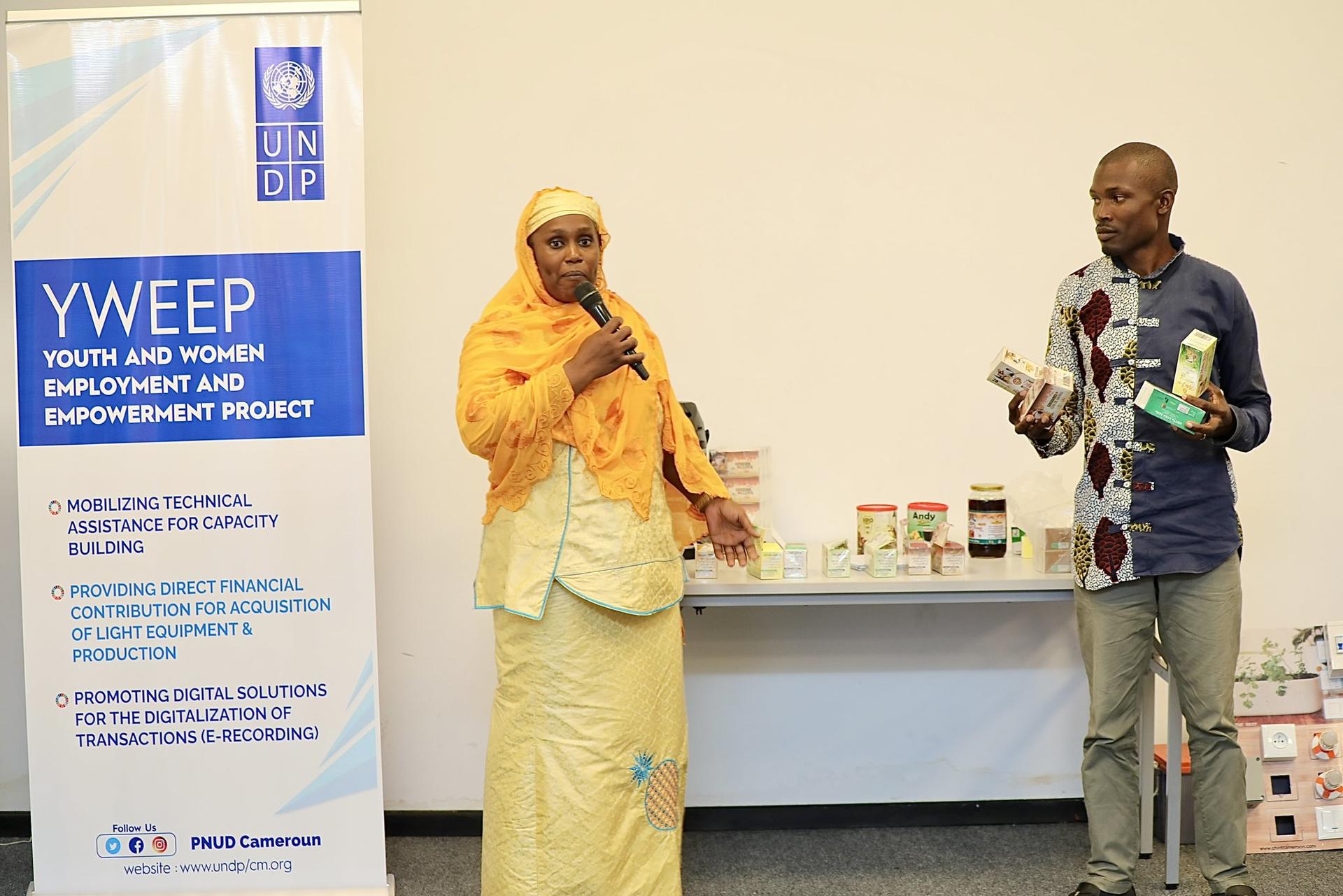 Woman in yellow dress and hijab and man in a dark shirt speak beside a banner at a product table.