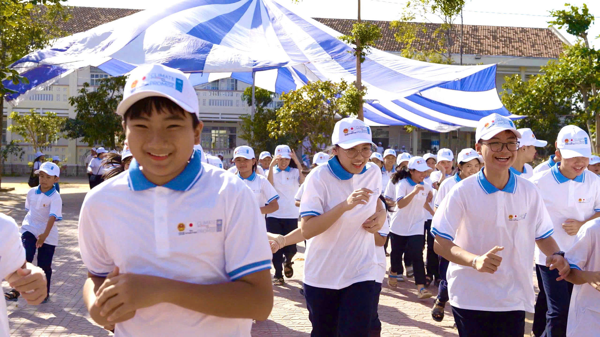 People in white shirts and caps gather at an outdoor event under blue-and-white tents.