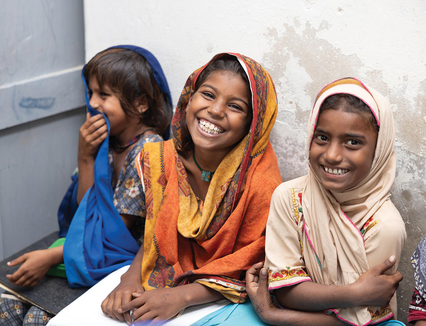 Three women in bright headscarves sit side by side against a pale wall.