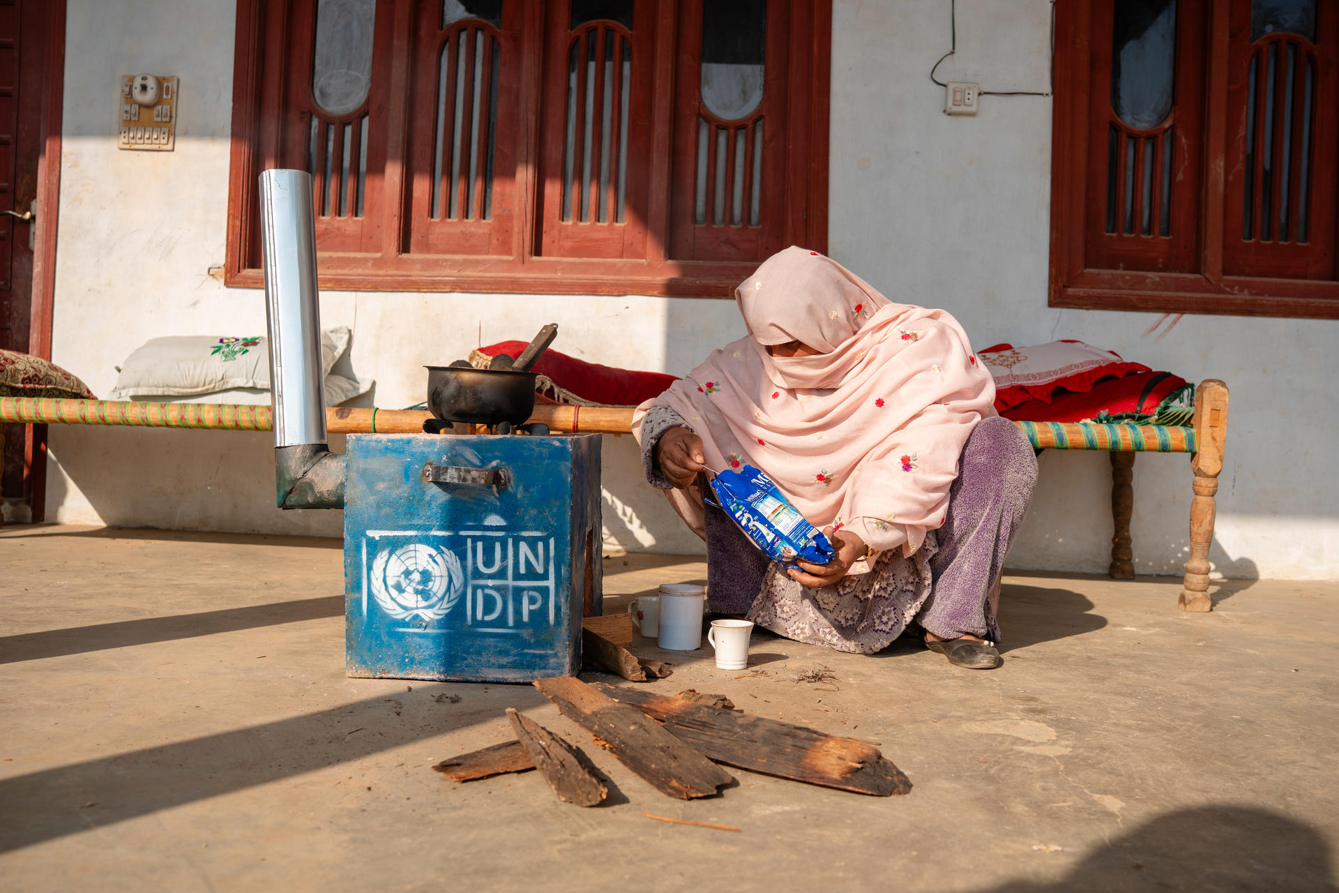 Woman with pink headscarf sits by a blue UNICEF bag and wood planks outside a building with red doors.