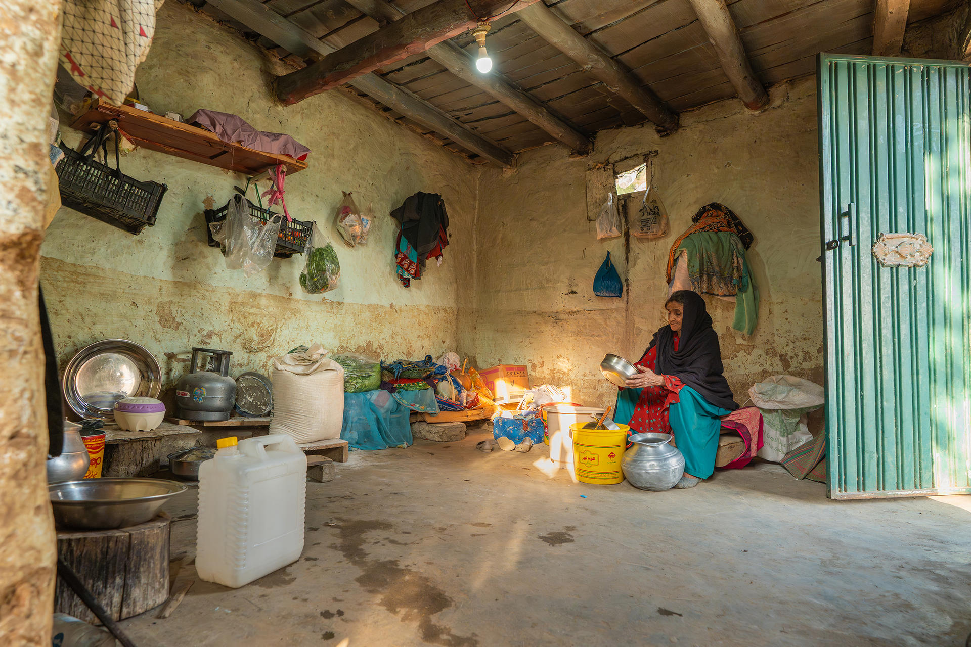 Rural kitchen with earthen walls, a woman at a small table, hanging pots, green corrugated door.