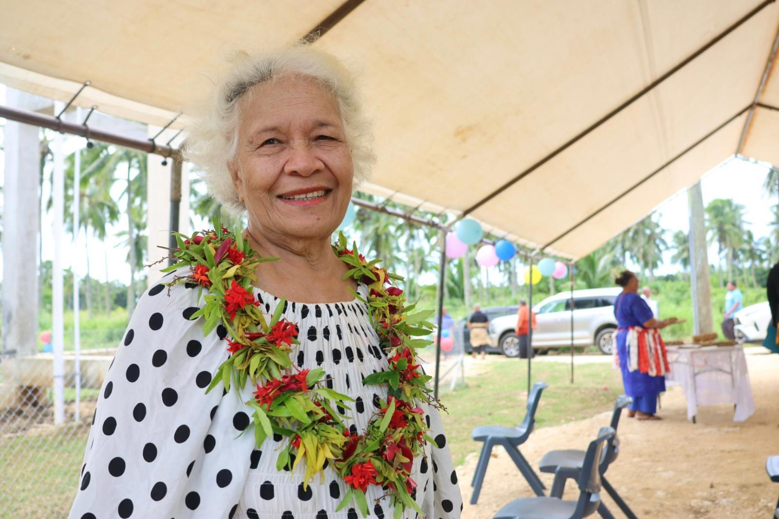 Person with blurred face wearing white polka-dot dress and a colorful lei under canopy outdoors.