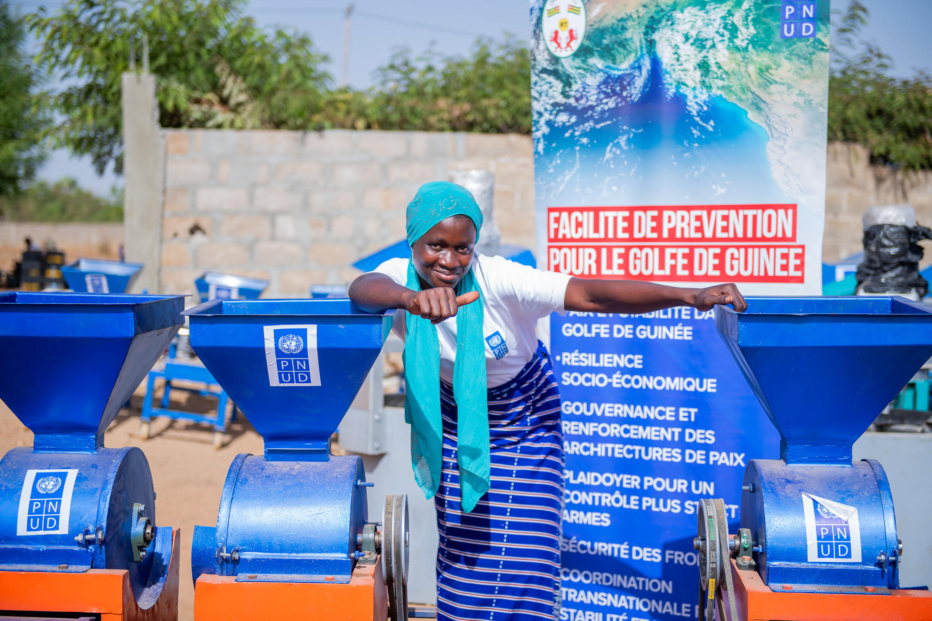 Outdoors, a person wearing a turquoise headscarf speaks beside blue machinery and a banner.