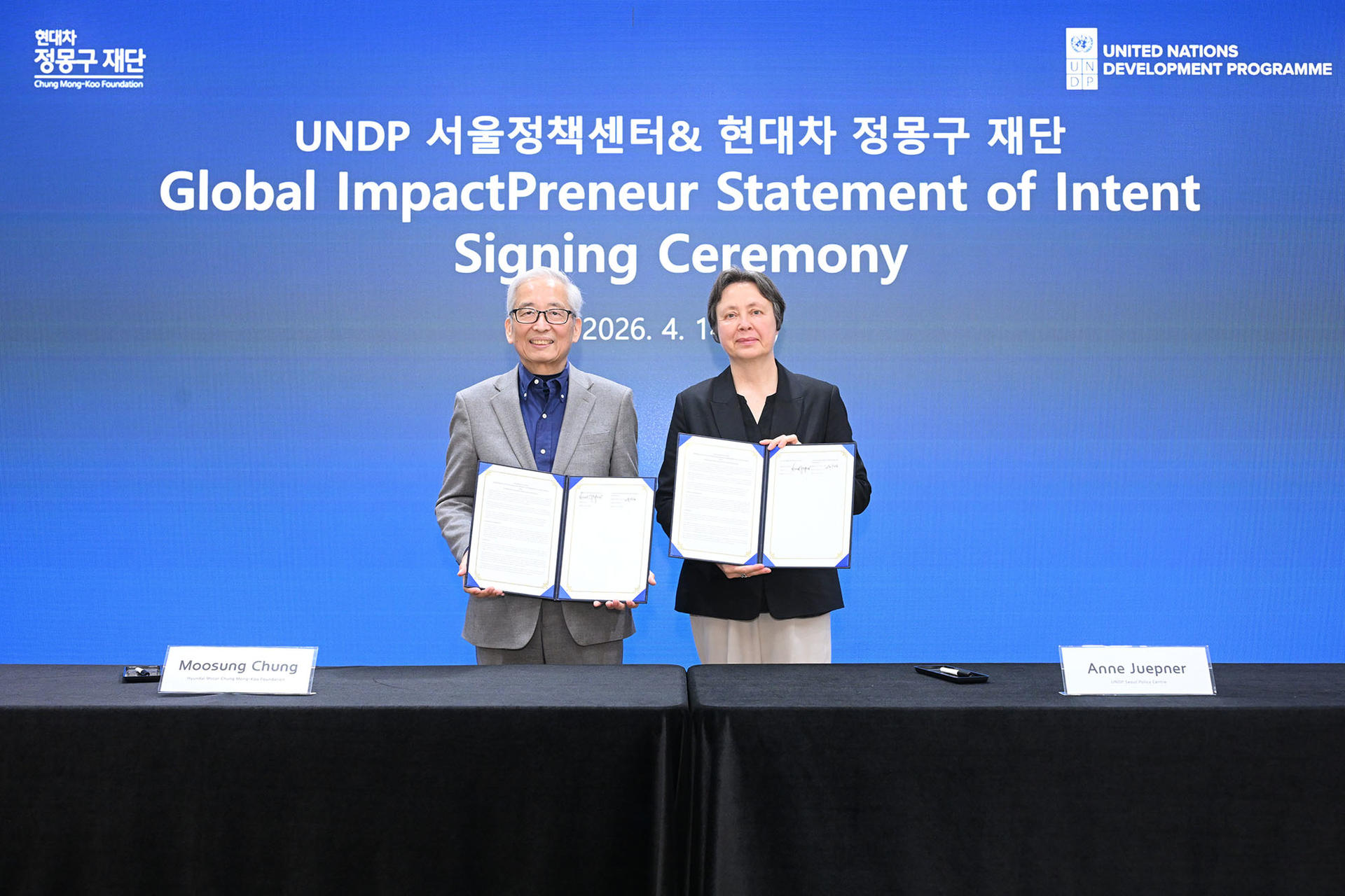 Photograph of two professionals in suits holding documents at a signing ceremony on a blue backdrop.