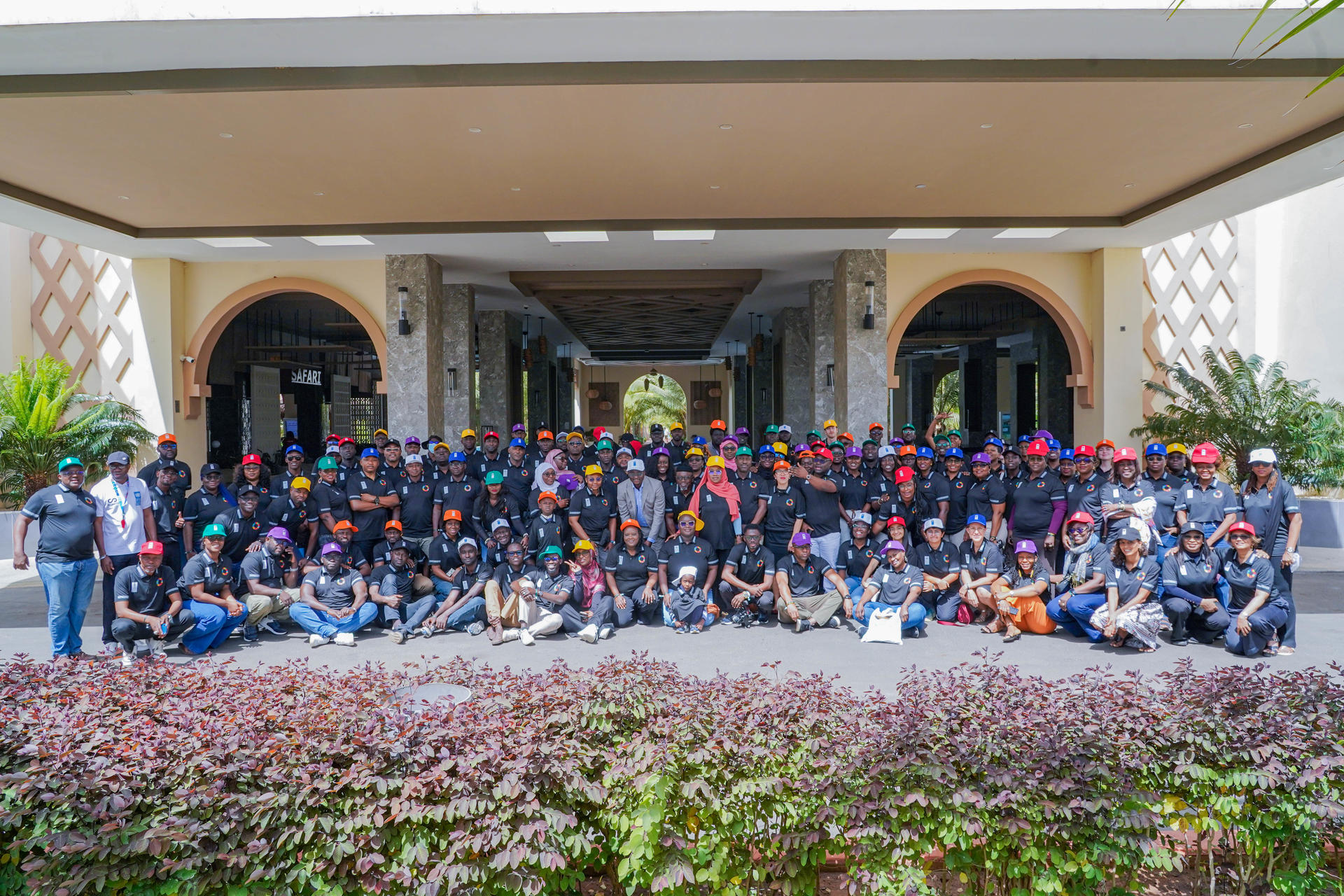 Large group of people posing outside a building with arches, columns, and a flower bed.