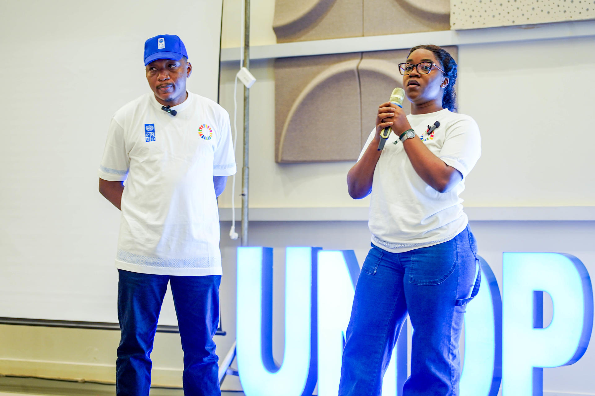 two UNDP staff (a man and a woman) on a stage with a glowing backdrop; one speaks into a microphone, the other stands beside.