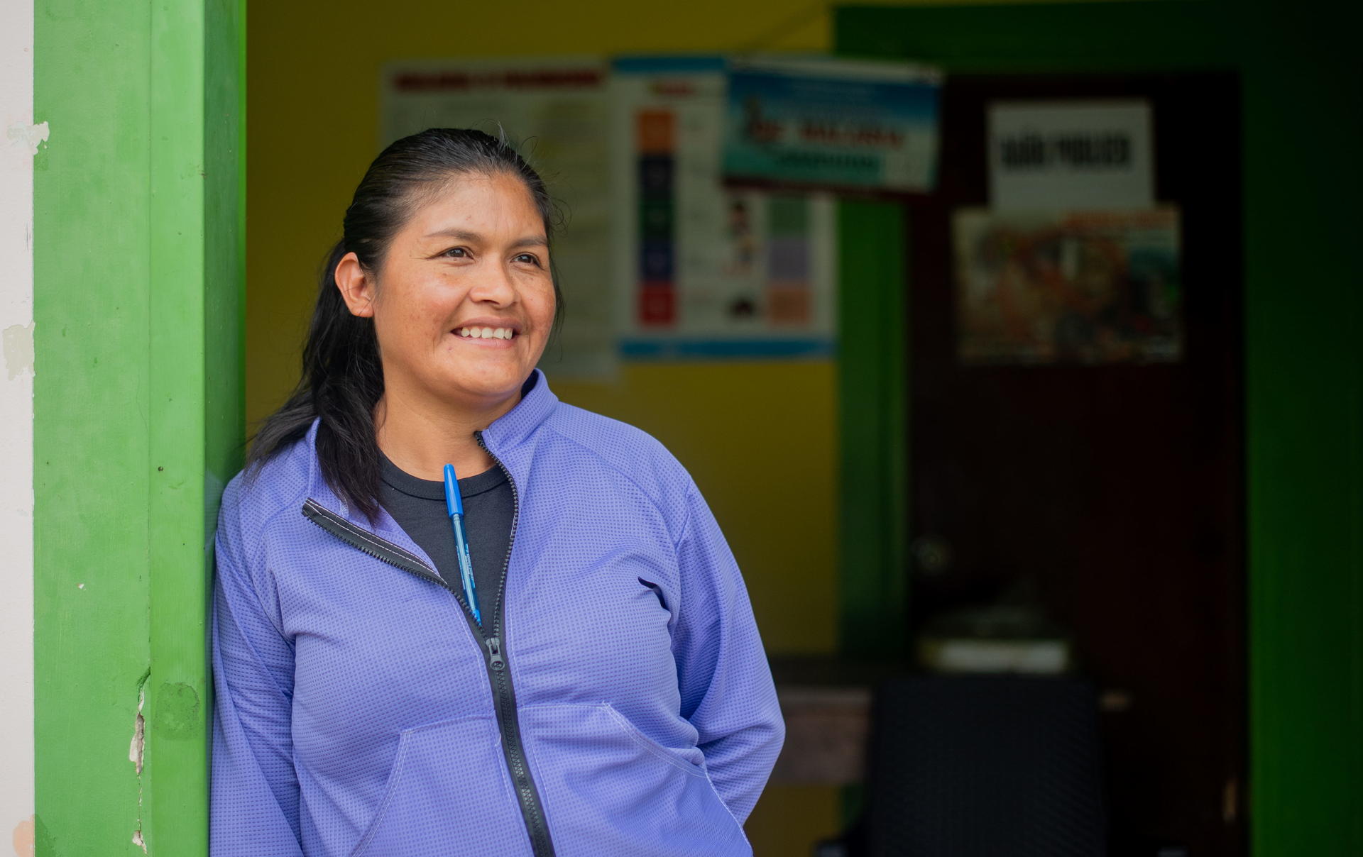 Photograph of a person in a blue jacket standing in a doorway; green wall and posters in the background, face blurred.