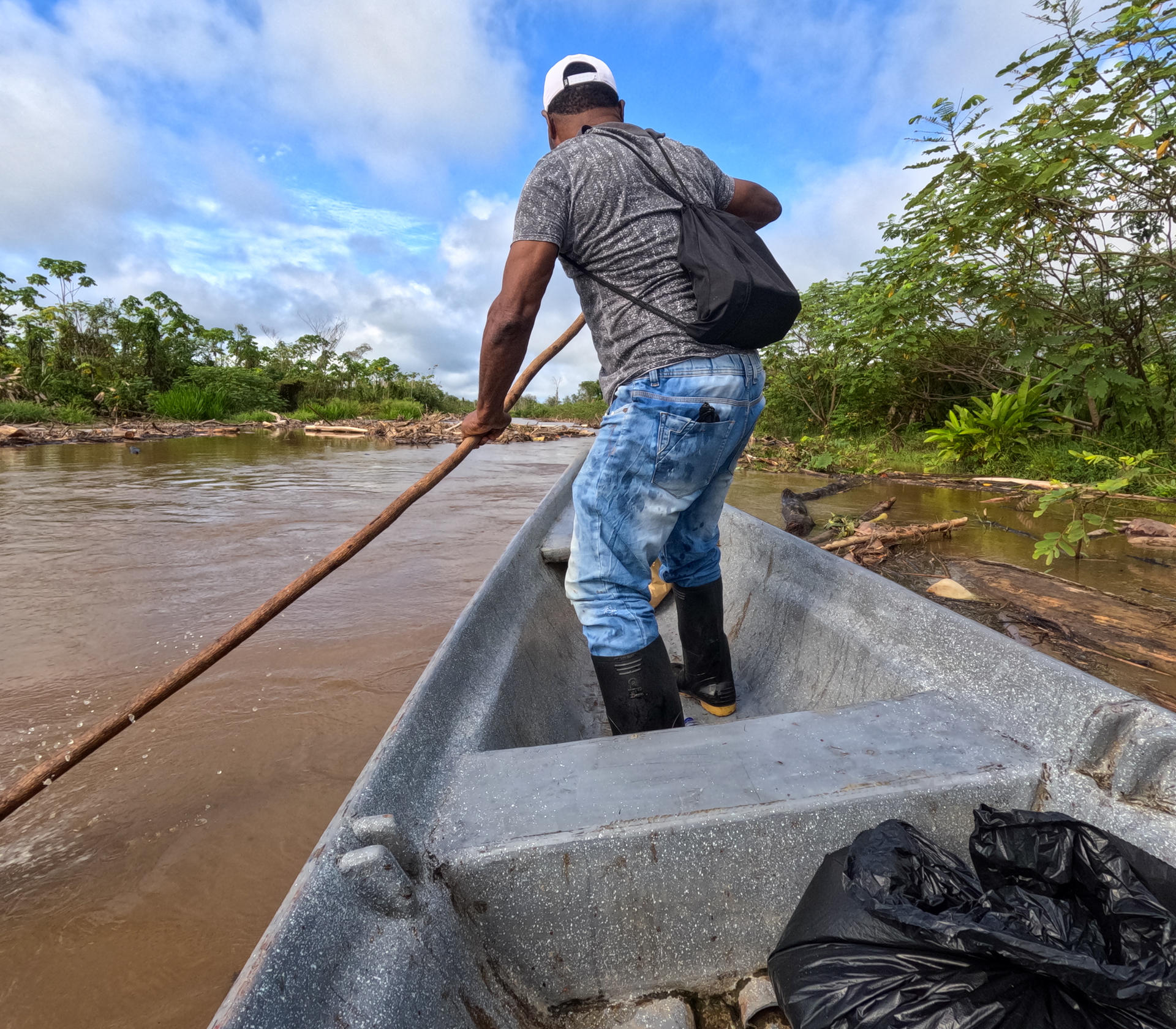 Man in jeans and boots steers a small aluminum boat on a muddy river with a long pole.