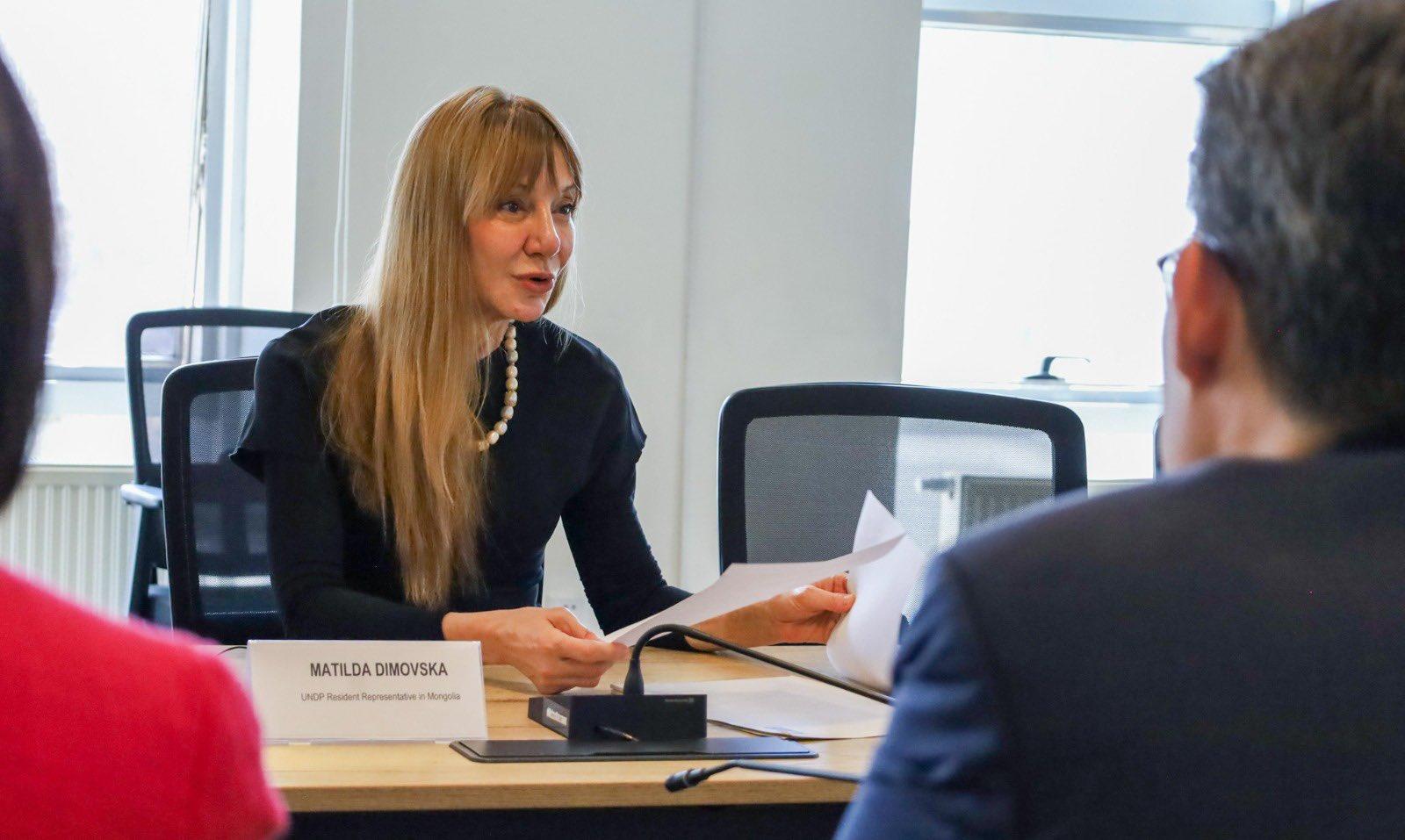 Blonde woman seated at a desk with a nameplate in a conference room, facing a man.