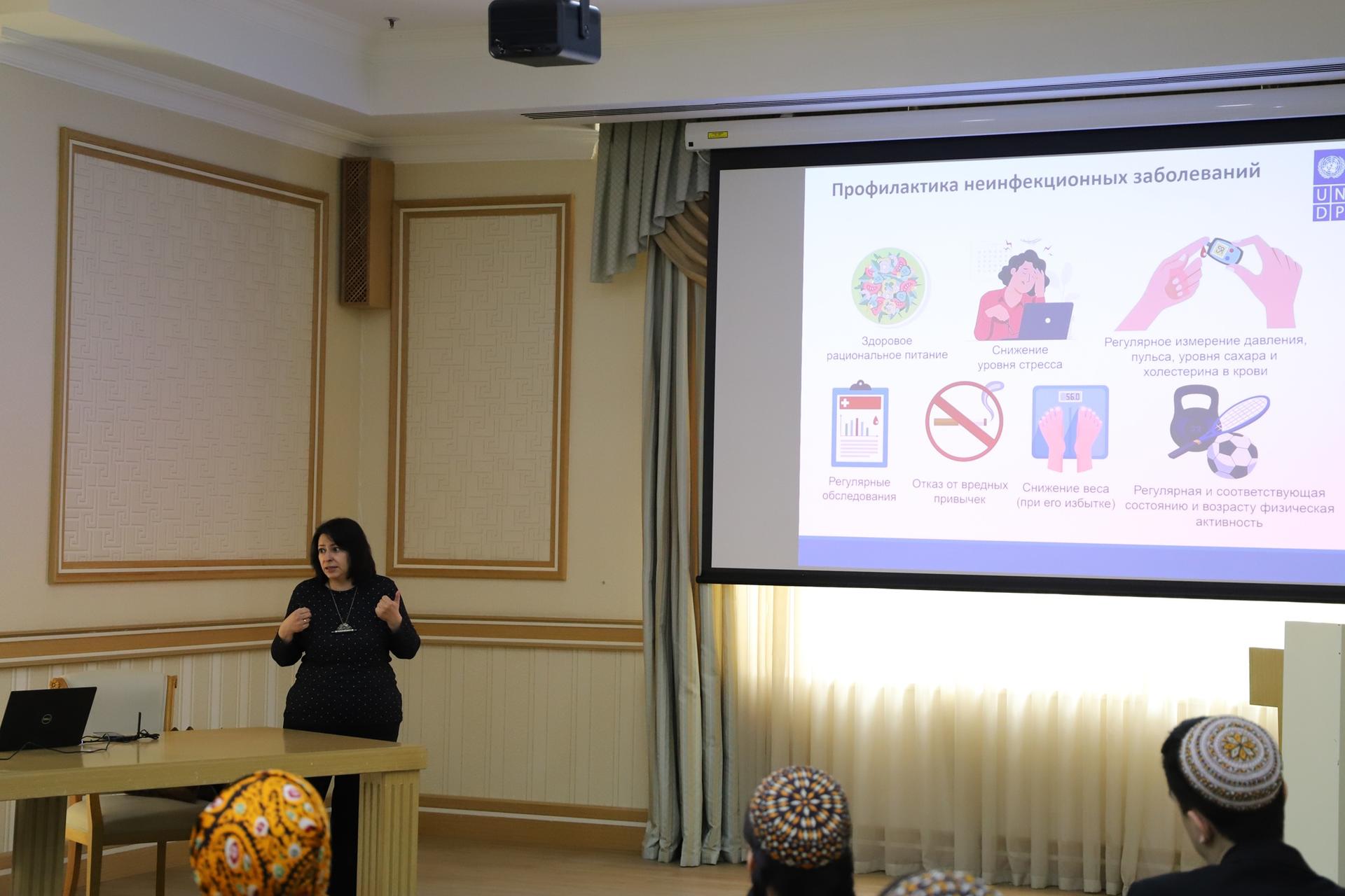 Female presenter in a beige conference room gives a talk; slide with colorful icons projected.