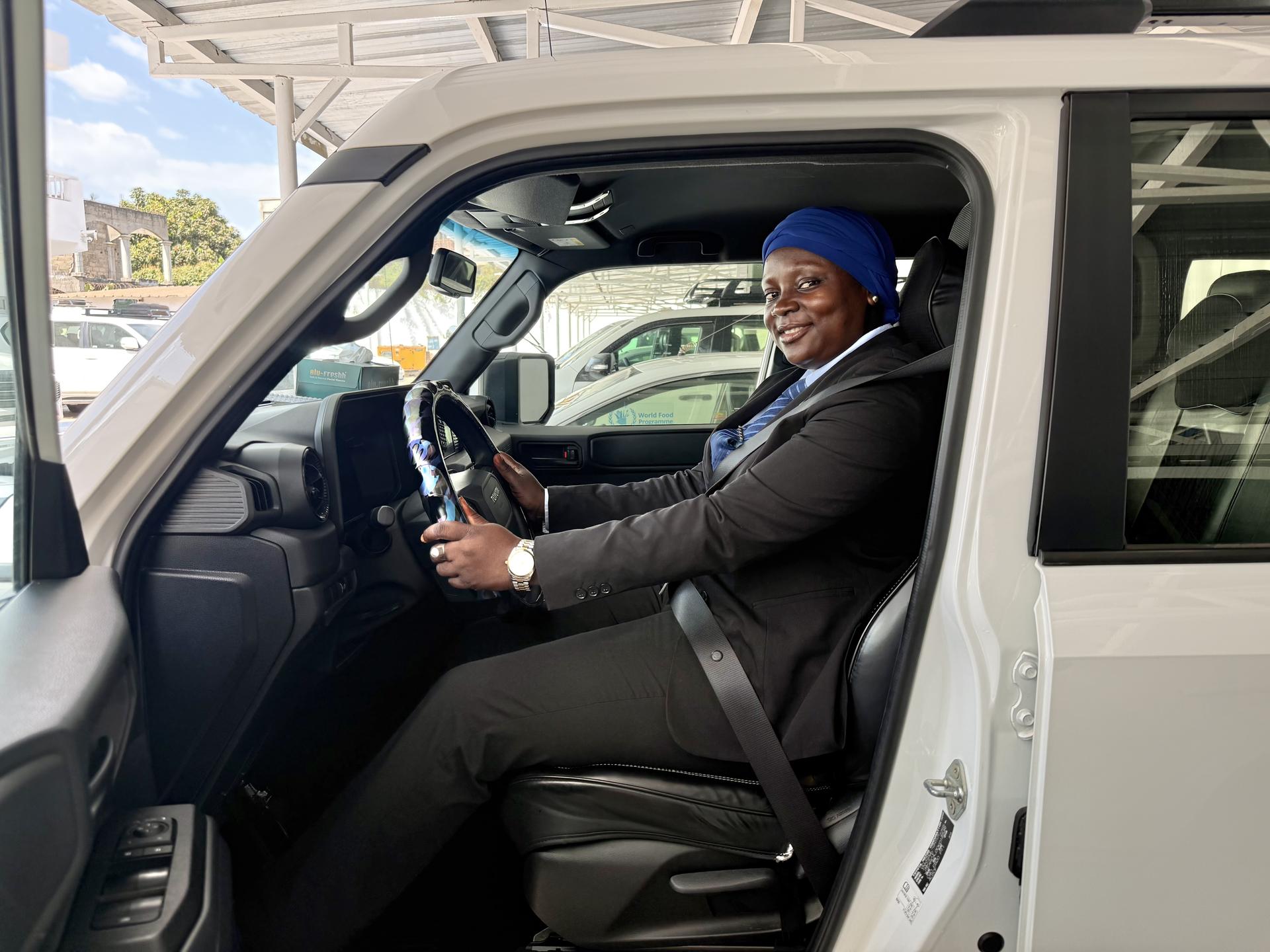 Photograph of a smiling woman driving a white van with an open door; blue headwrap.