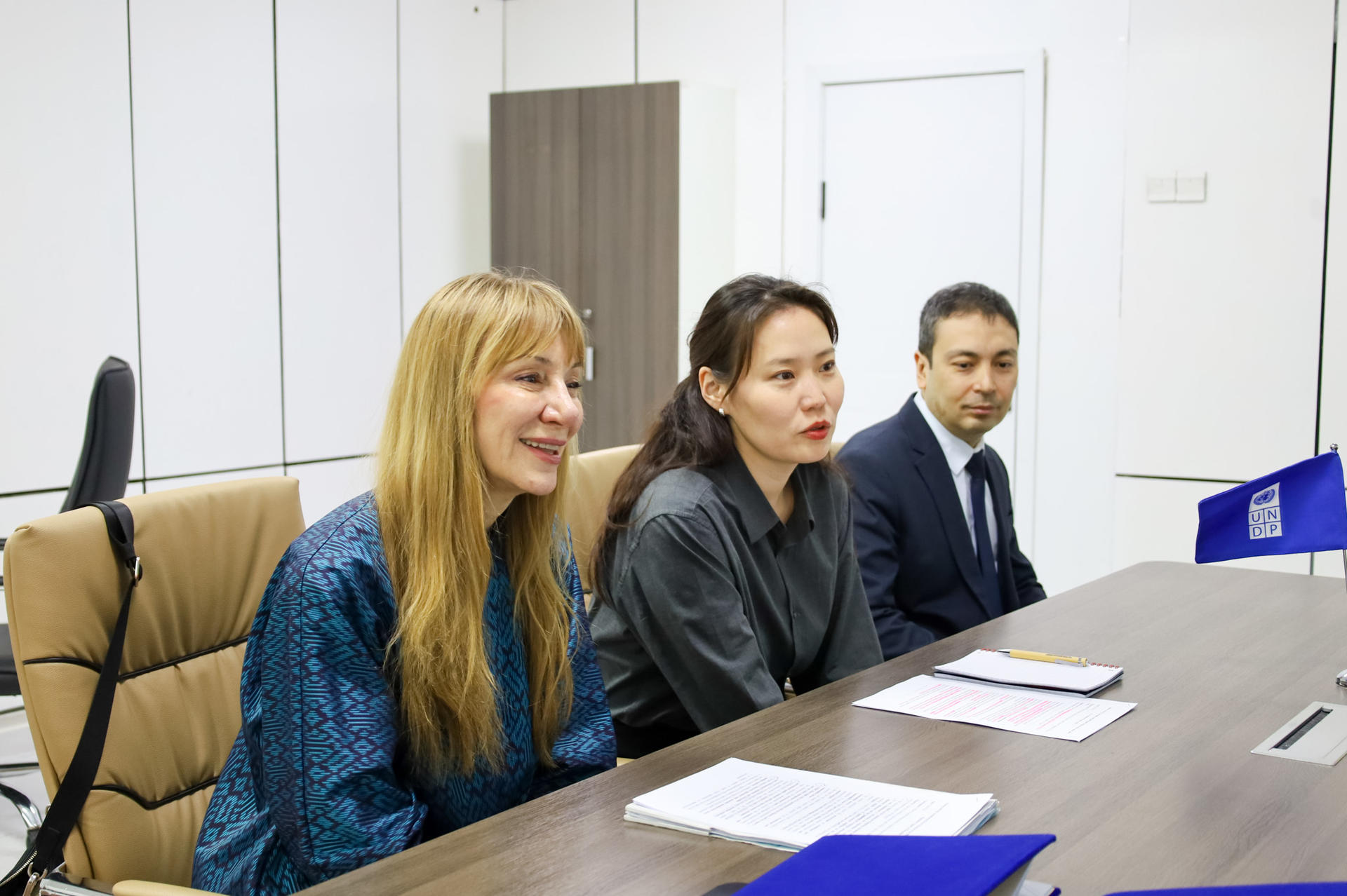 Three people in an office meeting sit at a long table with papers and blue folders.