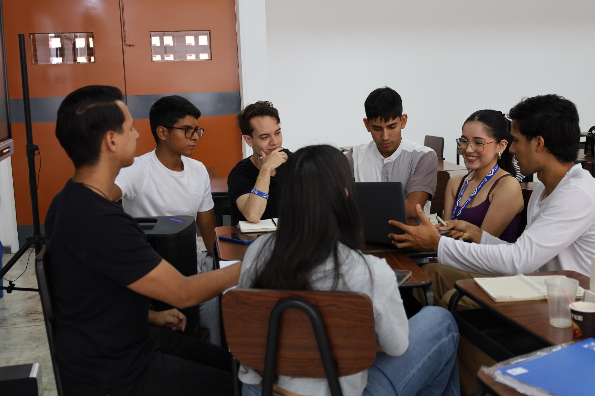 Group of students in a classroom, seated around a table and collaborating with papers and a laptop.