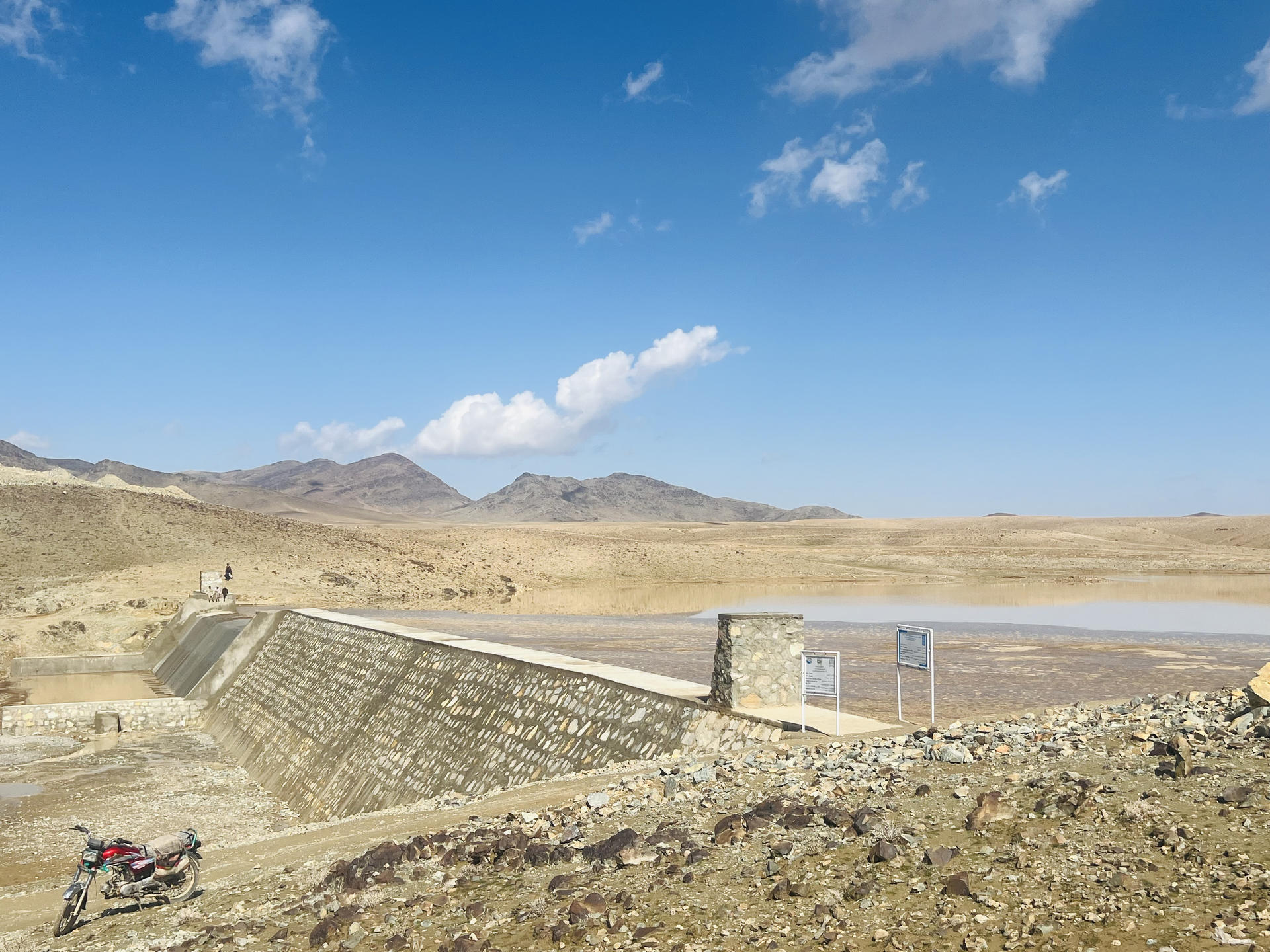 Desert landscape with a stone terraced wall along a slope, blue sky and distant mountains.